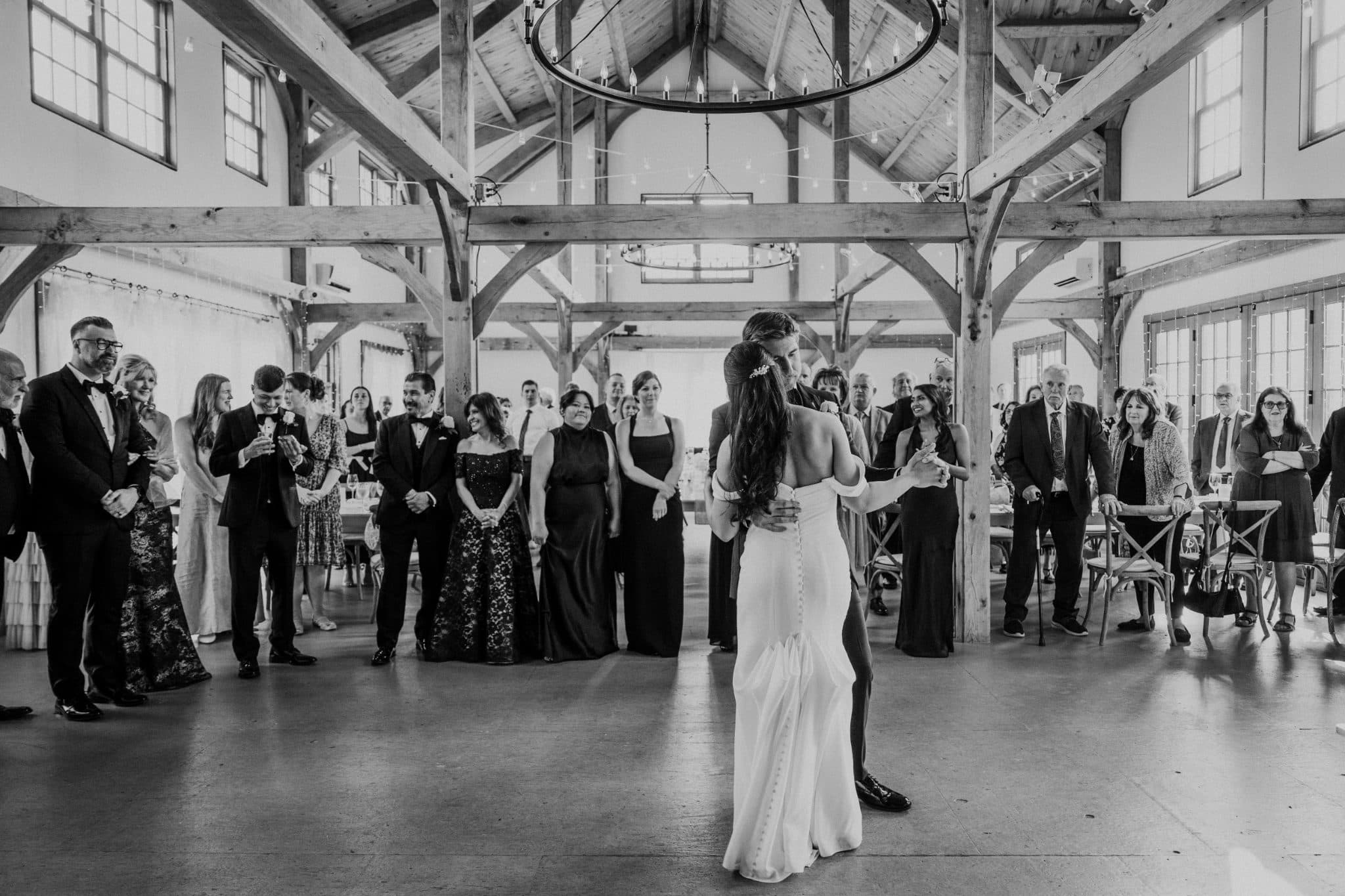 A bride and groom share their first dance in a rustic barn with wooden beams and a large chandelier.