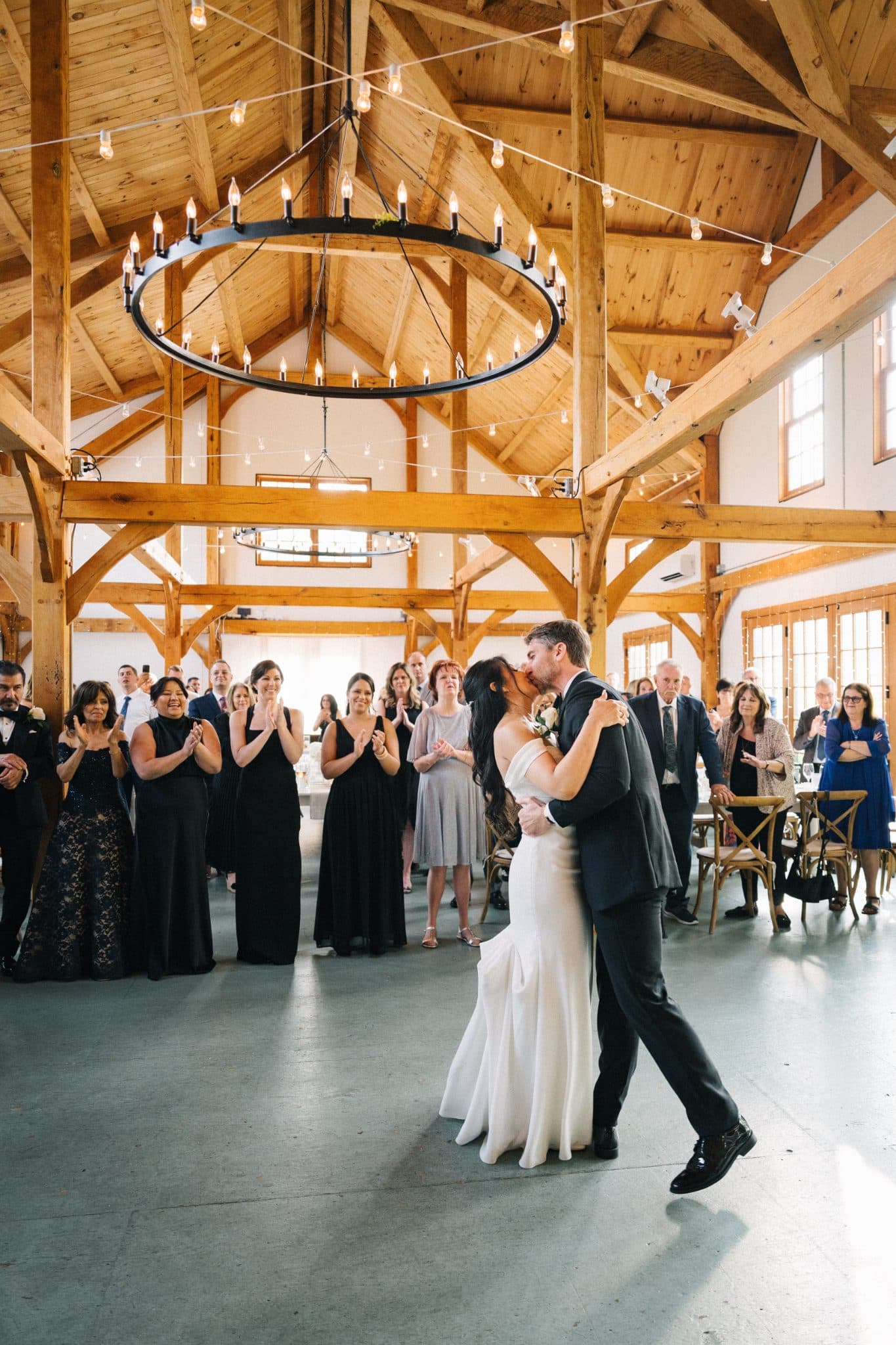 A bride and groom share a kiss during their first dance at a wedding reception in a barn with wooden beams and string lights.