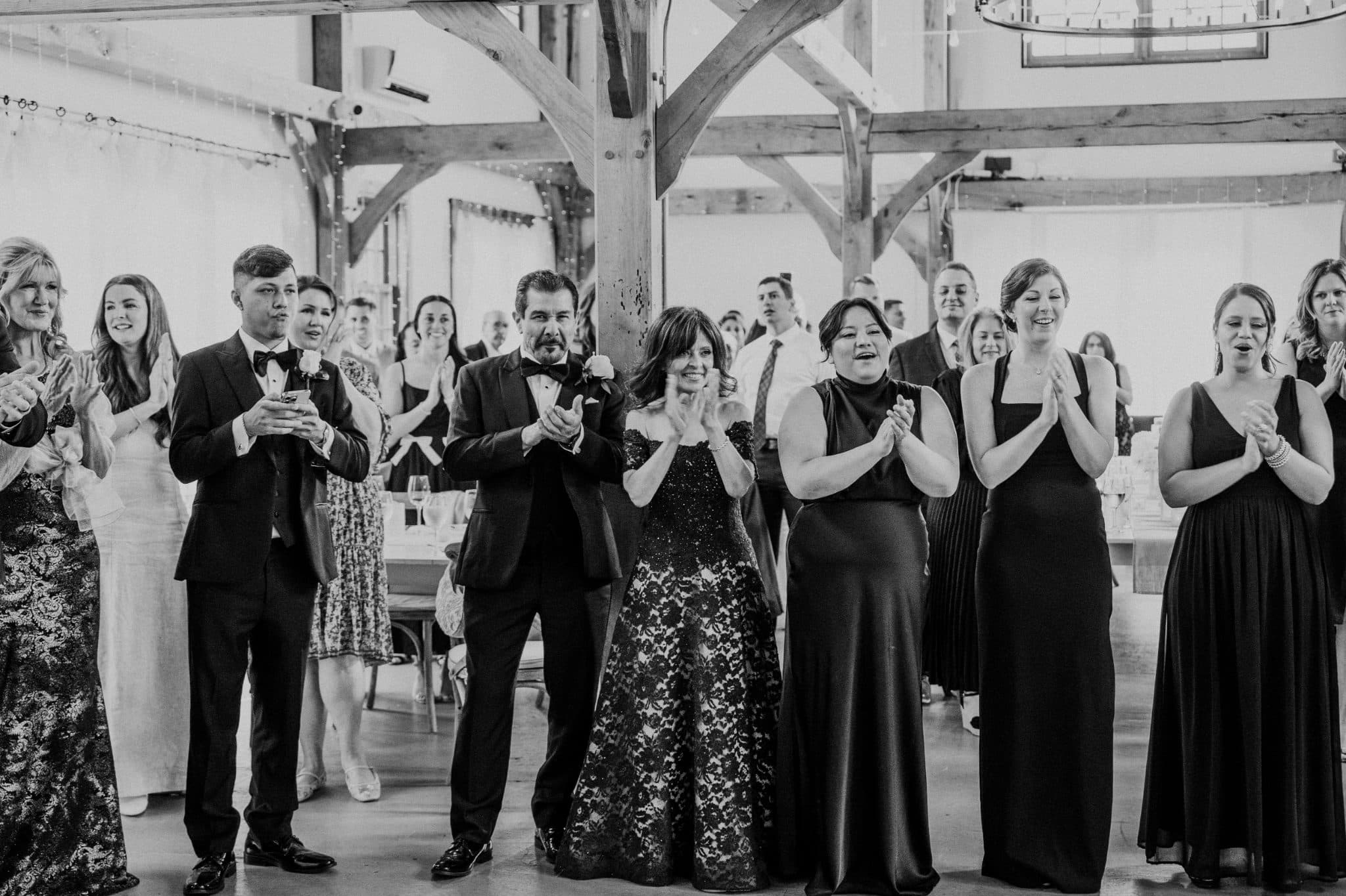 A group of people in formal attire claps in a barn-style venue with wooden beams.