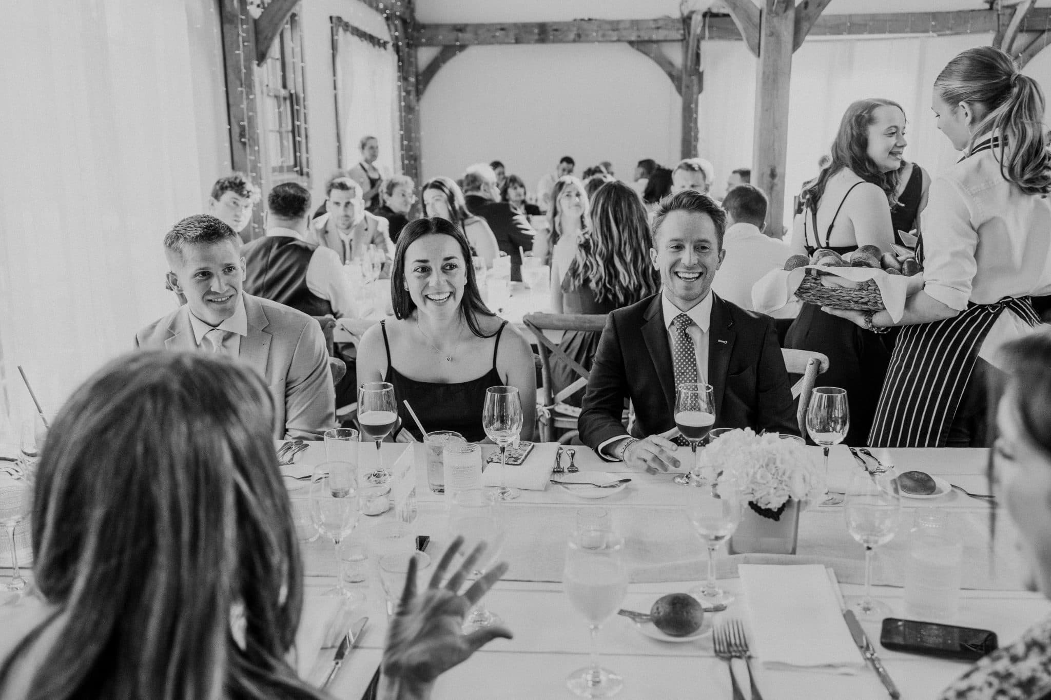 A black-and-white wedding reception scene with guests seated at tables, smiling, and a server holding a basket.
