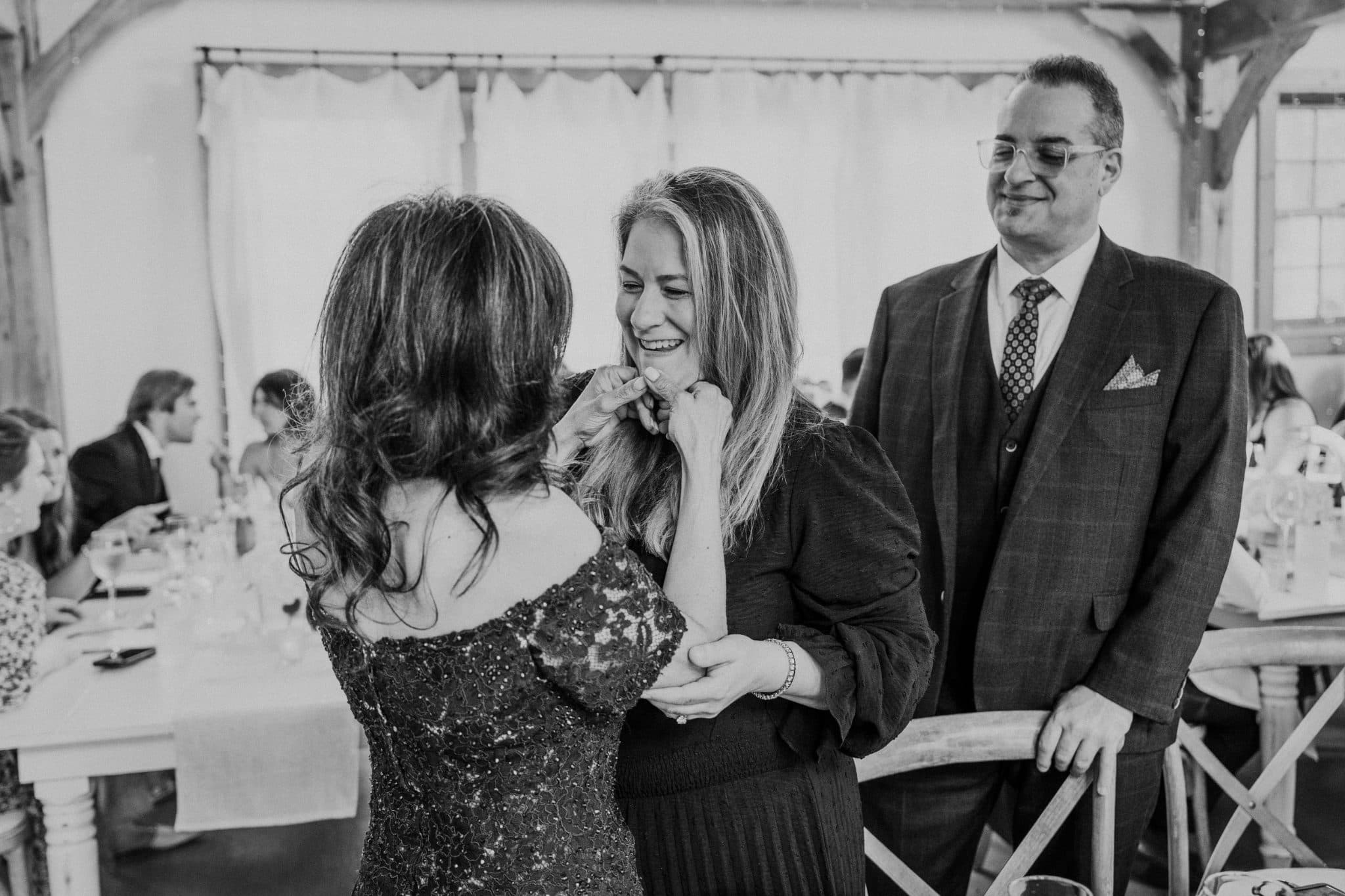 A woman in a sequined dress adjusts another woman's necklace while a man in a suit stands nearby in a room with tables and chairs.