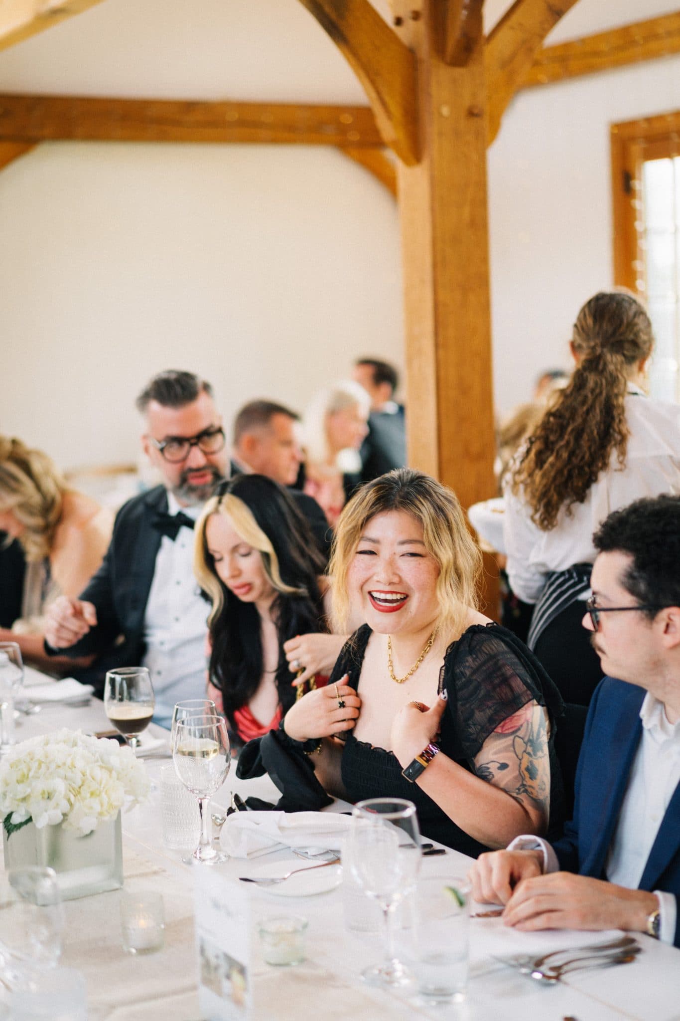 A woman with tattoos laughs at a table set for a formal event, surrounded by other guests.