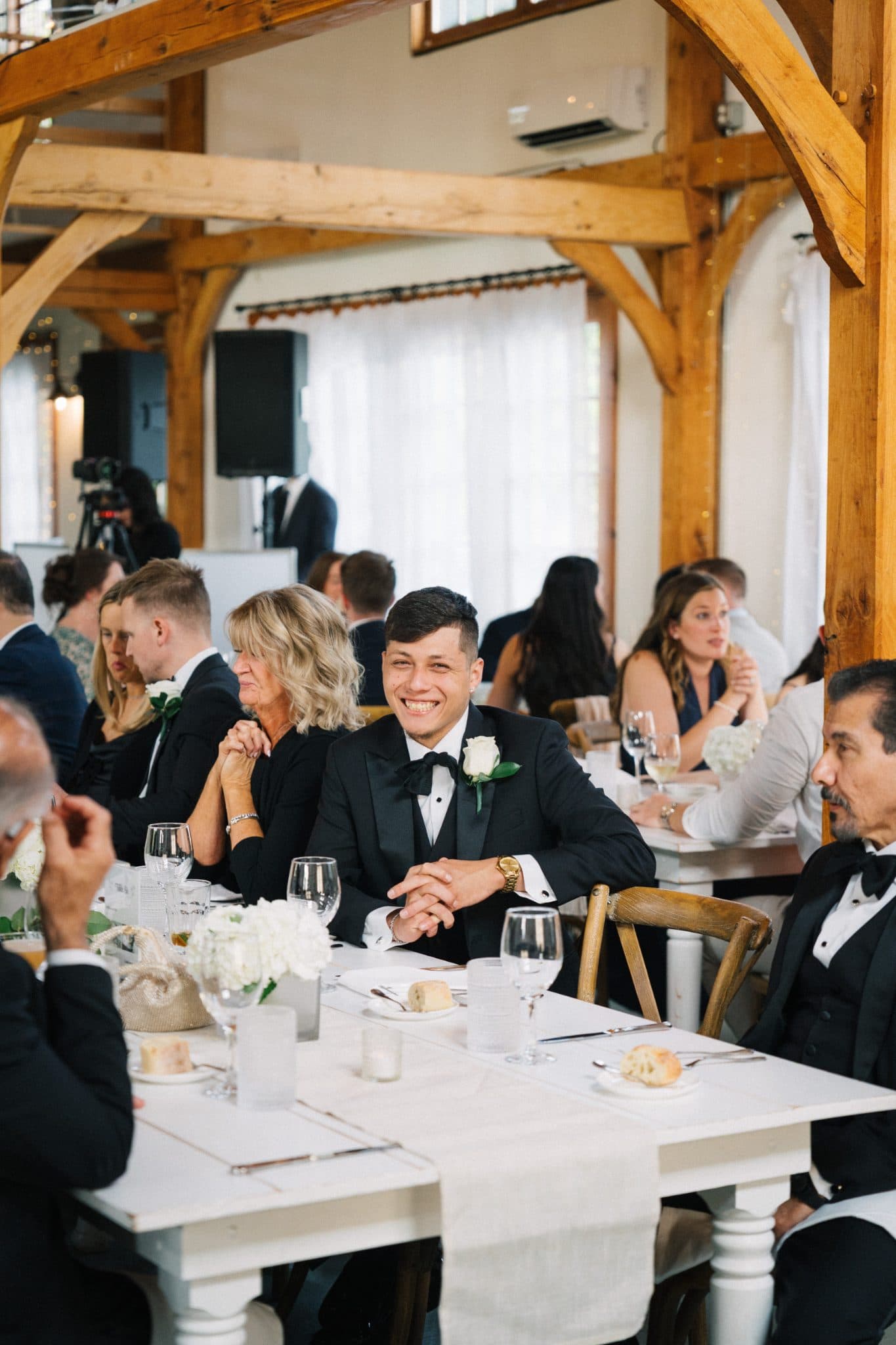 A man in a tuxedo smiles at the camera while seated at a table with other guests in a wooden-framed room.