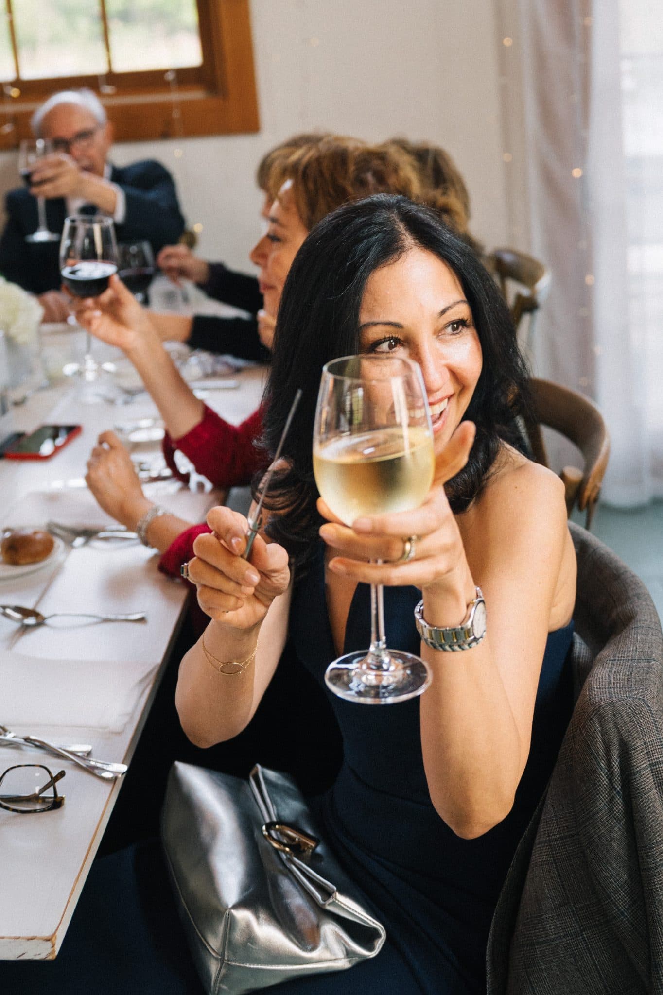 A woman in a black dress holds a glass of white wine, smiling at the camera, with others seated at a long table in the background.