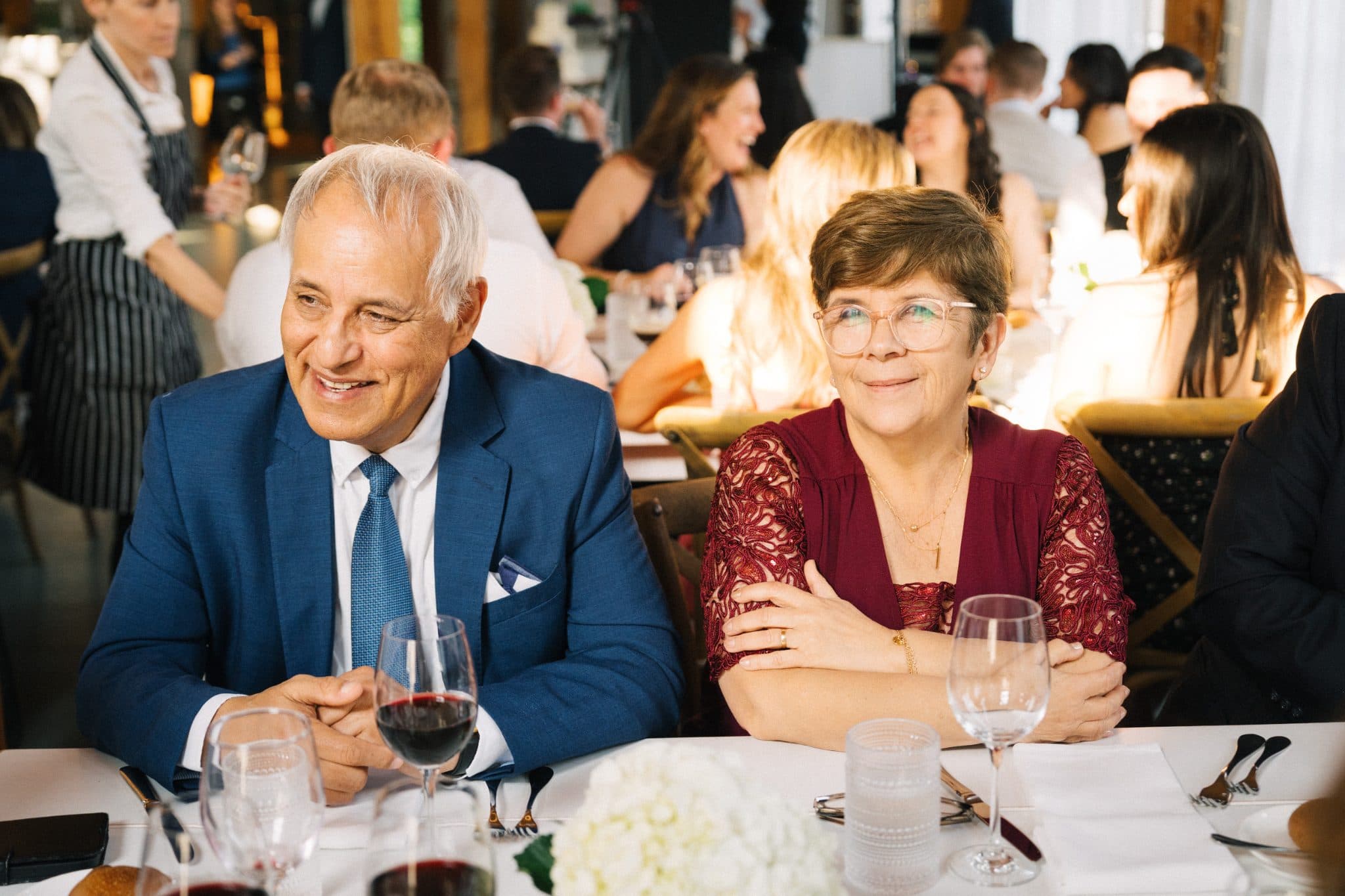 A man in a blue suit and a woman in a red patterned top sit at a table with wine glasses, smiling and looking at each other in a restaurant setting.