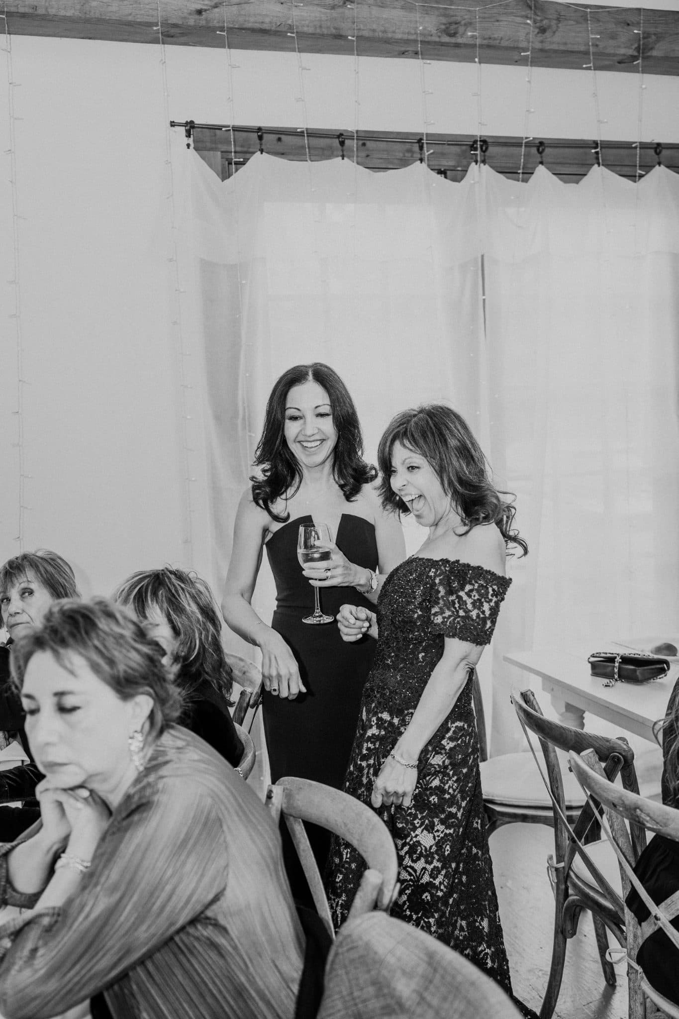 Two women in elegant dresses stand laughing, one holding a wine glass, surrounded by seated guests in a tented setting.