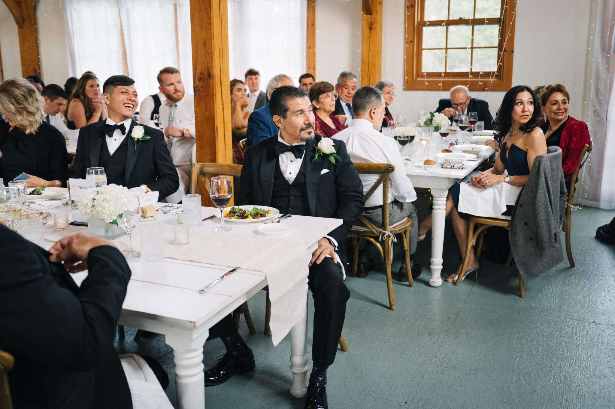 A wedding reception with guests in formal attire seated at white tables with white tablecloths and floral centerpieces.