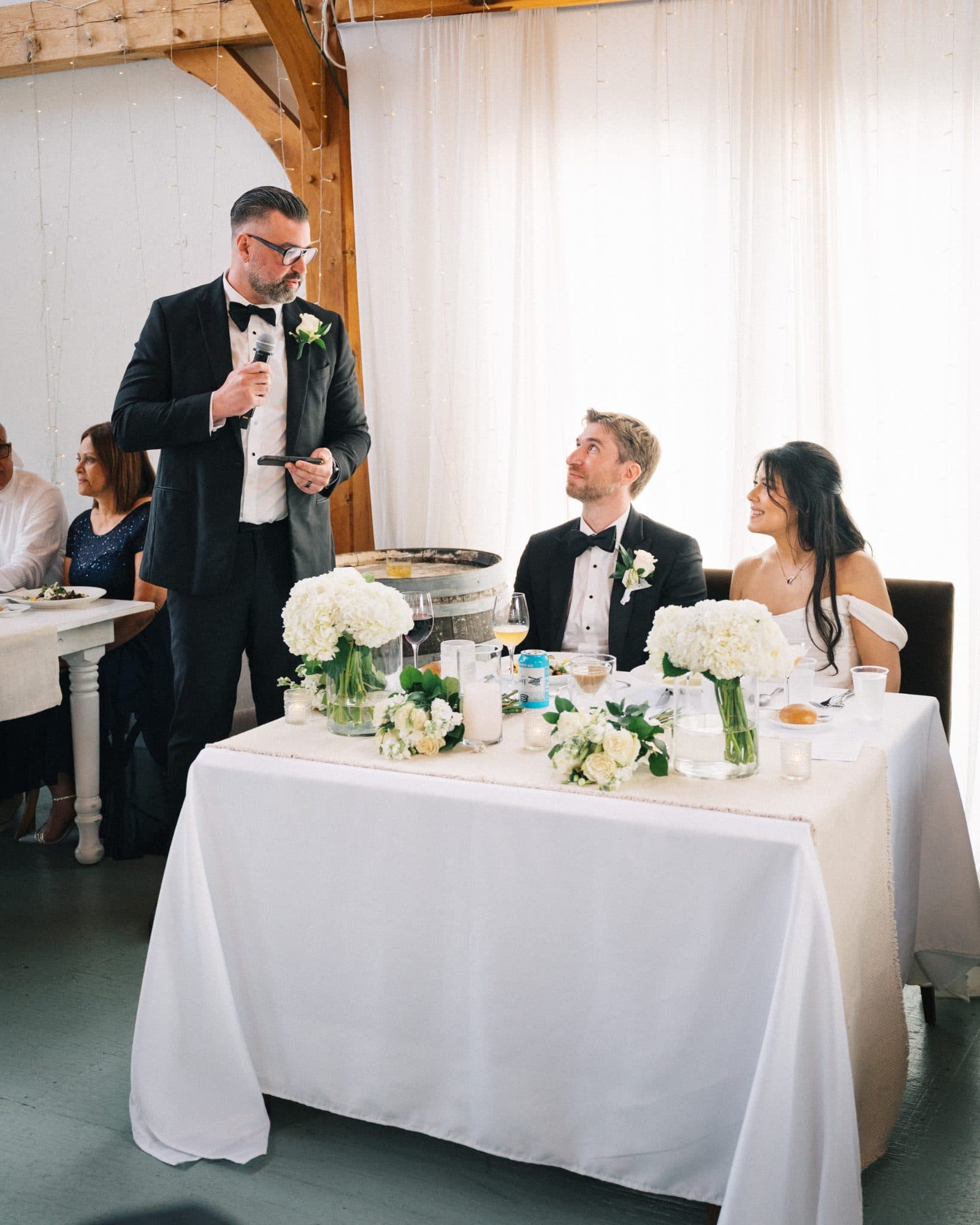 A man in a suit stands at a podium addressing a seated couple at a decorated table with white flowers and candles.