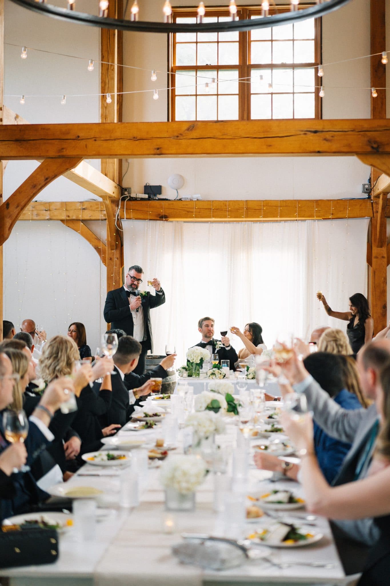A wedding reception with guests seated at a long table, raising glasses in a toast under a large circular chandelier.