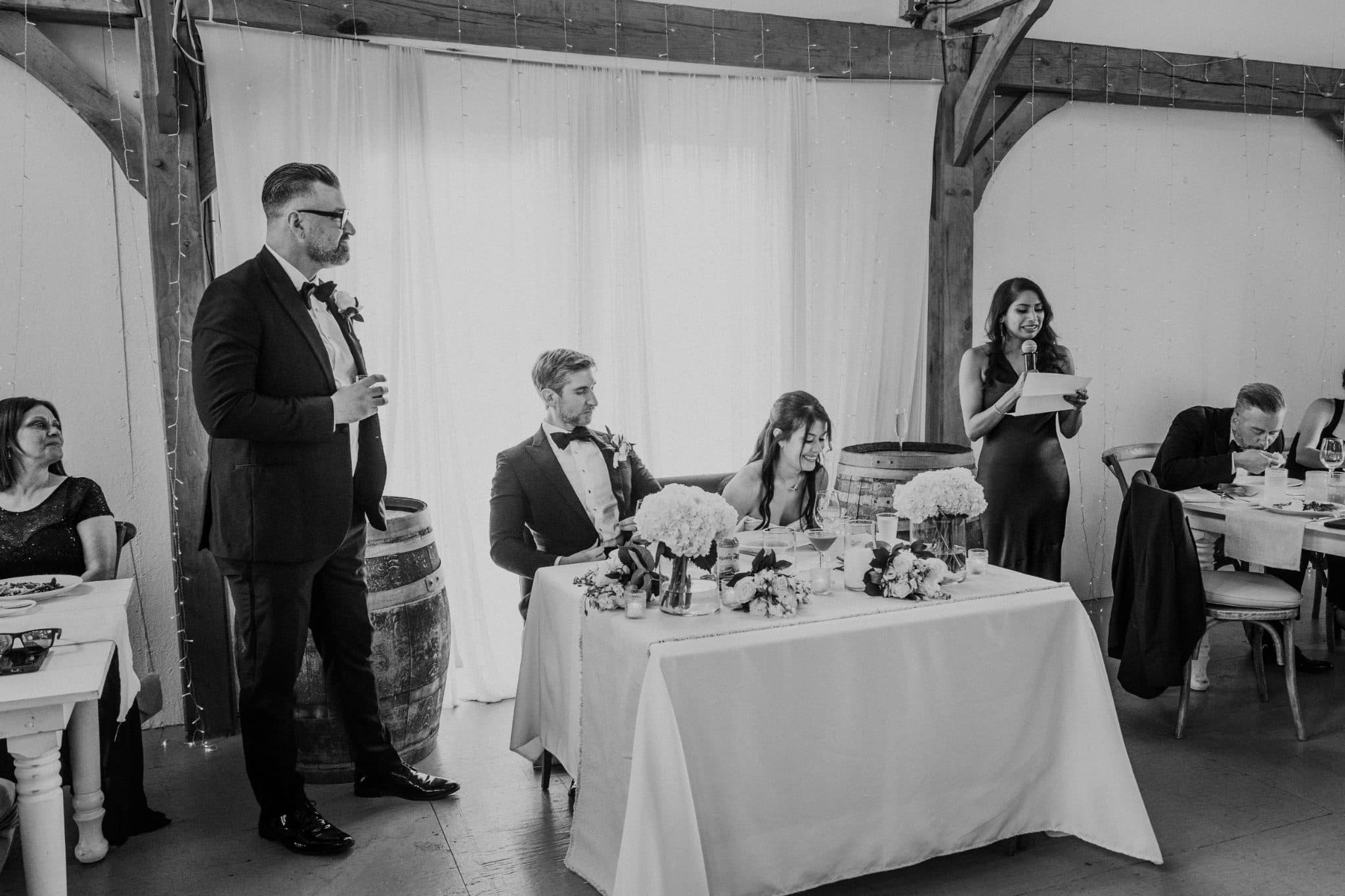 A man in a suit stands to the left, addressing a seated couple at a table adorned with flowers and wine glasses.