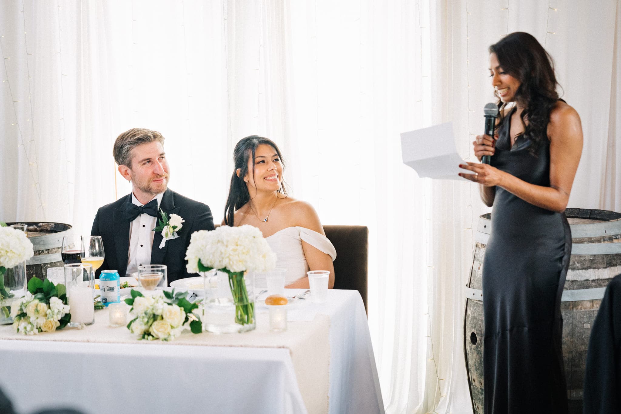 A woman in a black dress holds a microphone and reads from a paper while a couple sits at a table adorned with white flowers and wine glasses.