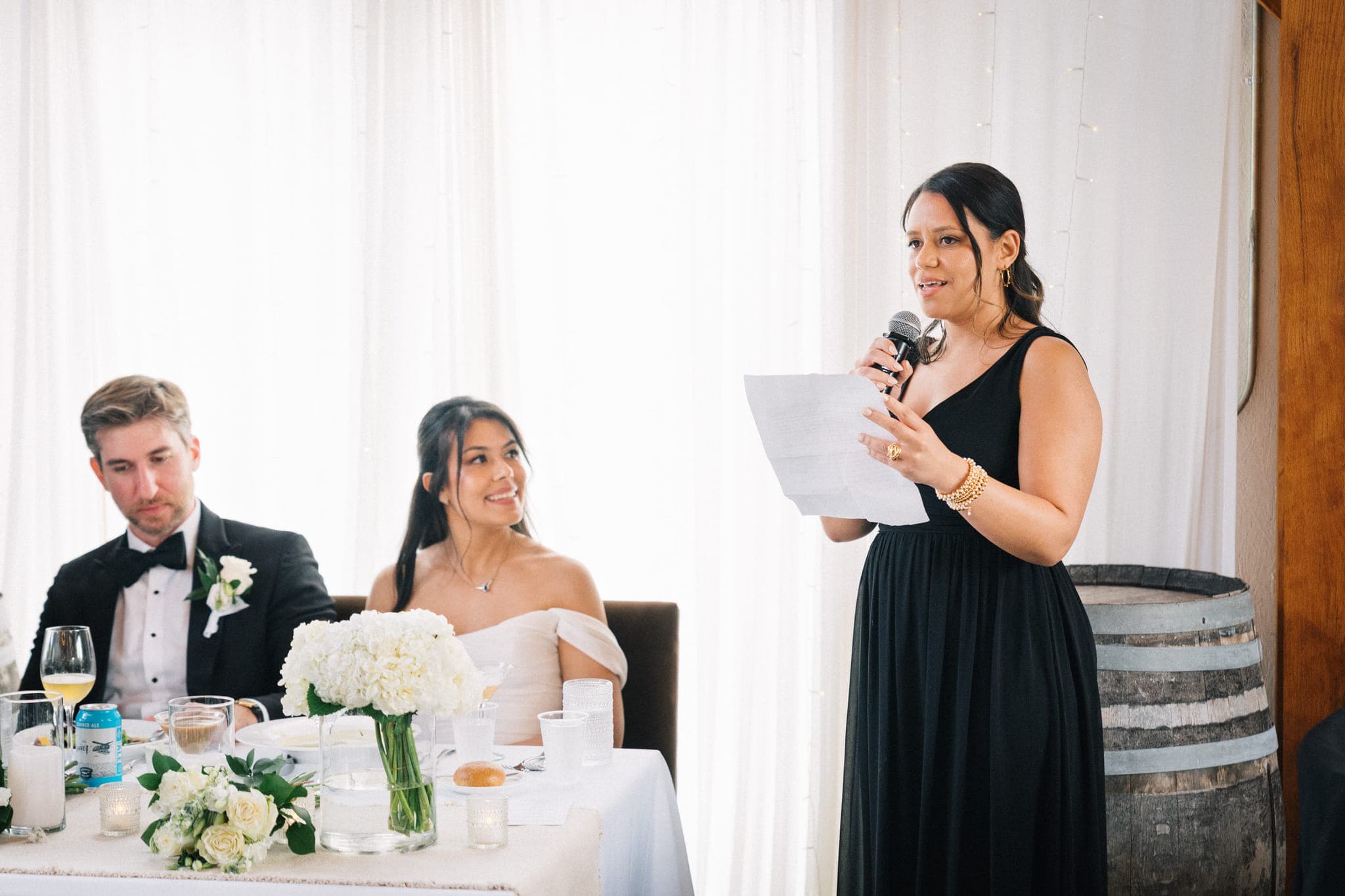 A woman in a black dress holds a microphone and paper, standing beside a table with a bride and groom seated, decorated with flowers and candles.
