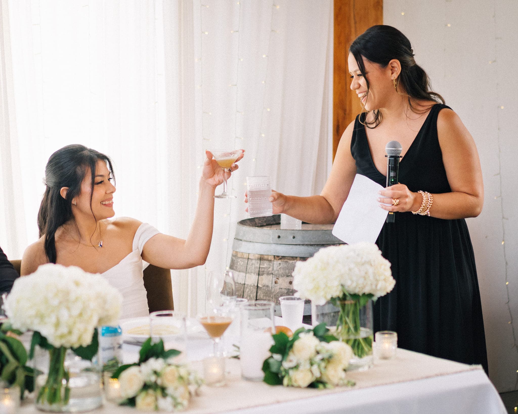 A woman in a black dress holds a microphone and a glass, toasting another woman in a white dress at a table adorned with white flowers and candles.