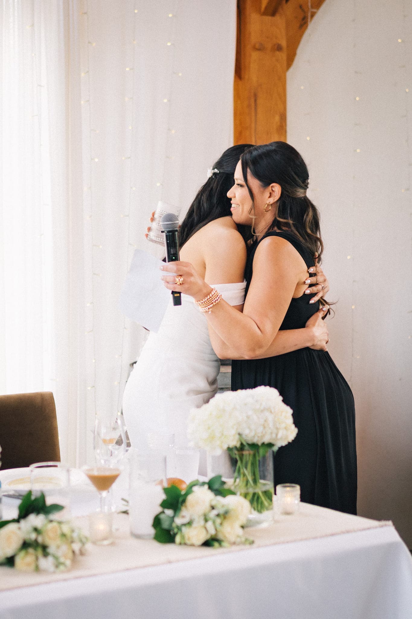 A bride in white embraces a woman in black, holding a microphone and paper, with a table adorned with flowers and drinks in the foreground.