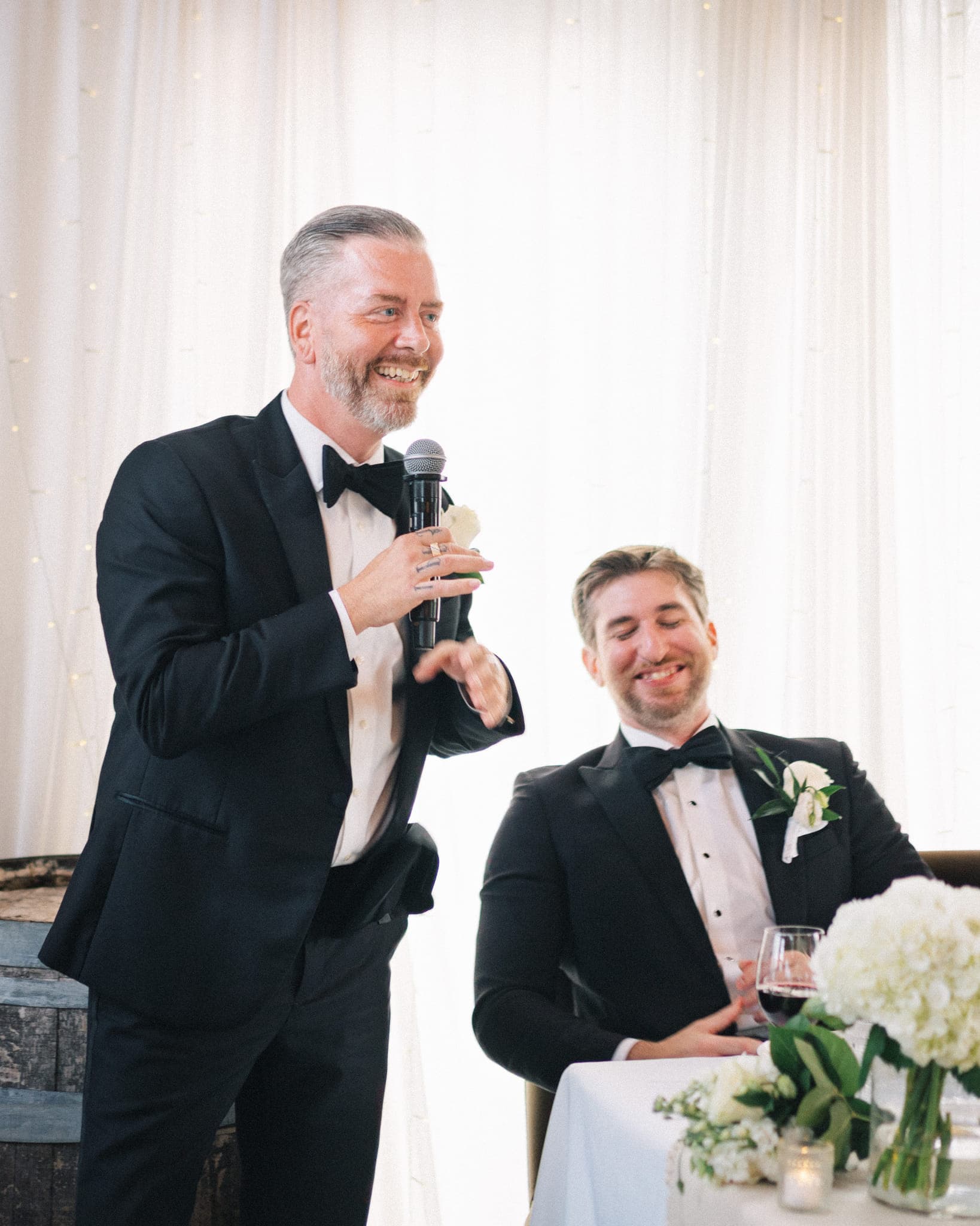 A man in a tuxedo holds a microphone, smiling, while another man in a tuxedo sits at a table with a white flower arrangement.