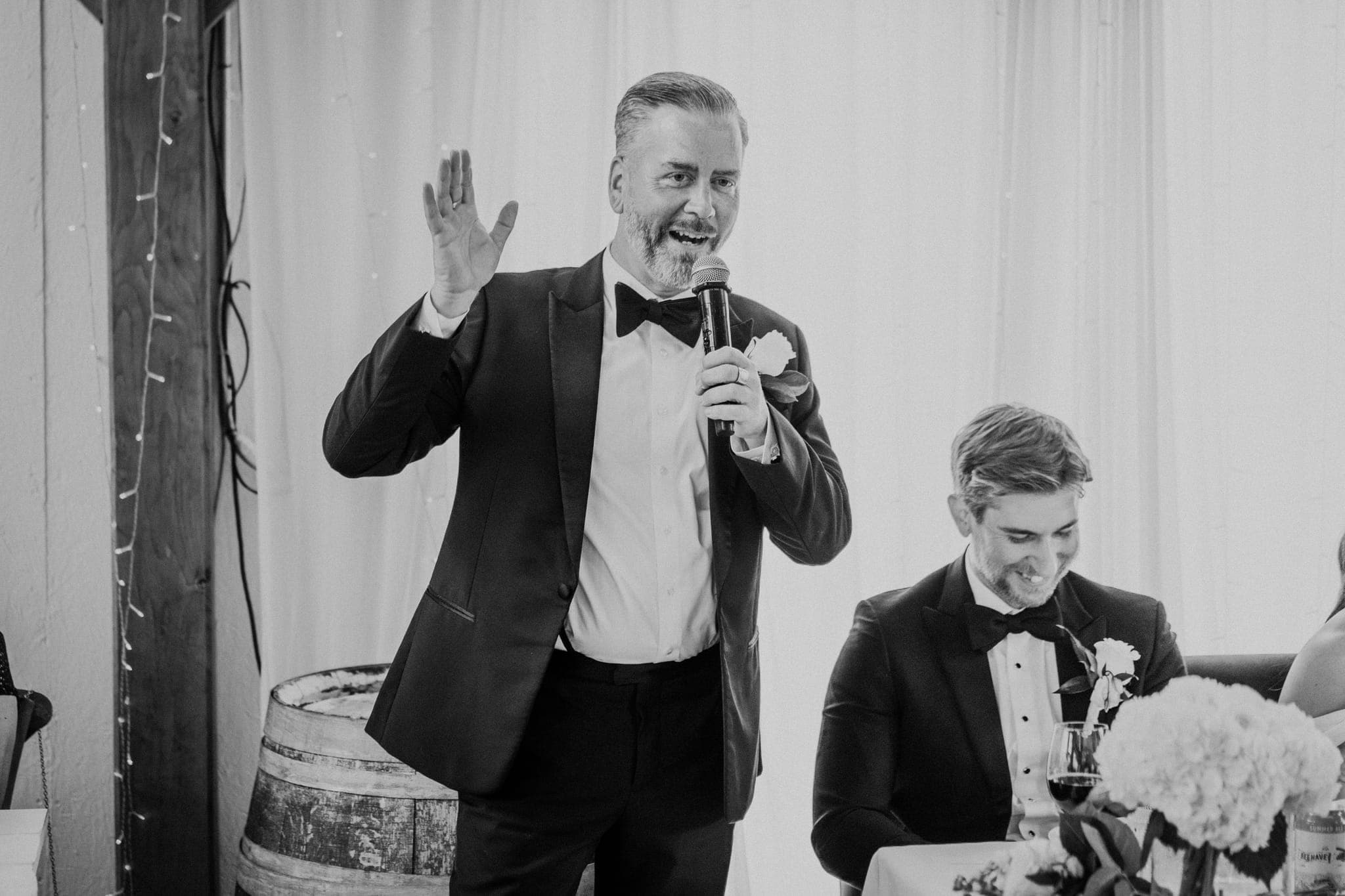 A man in a tuxedo stands holding a microphone, gesturing with his other hand, while another man in a tuxedo sits at a table with a glass of wine and a bouquet…