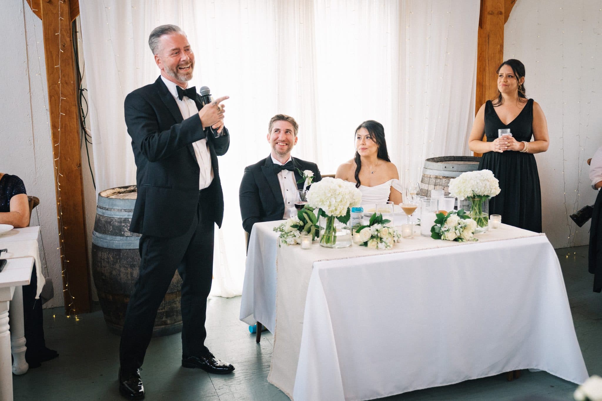 A man in a tuxedo stands at a podium, gesturing, while a couple and a woman in a black dress sit at a table adorned with white flowers and candles.