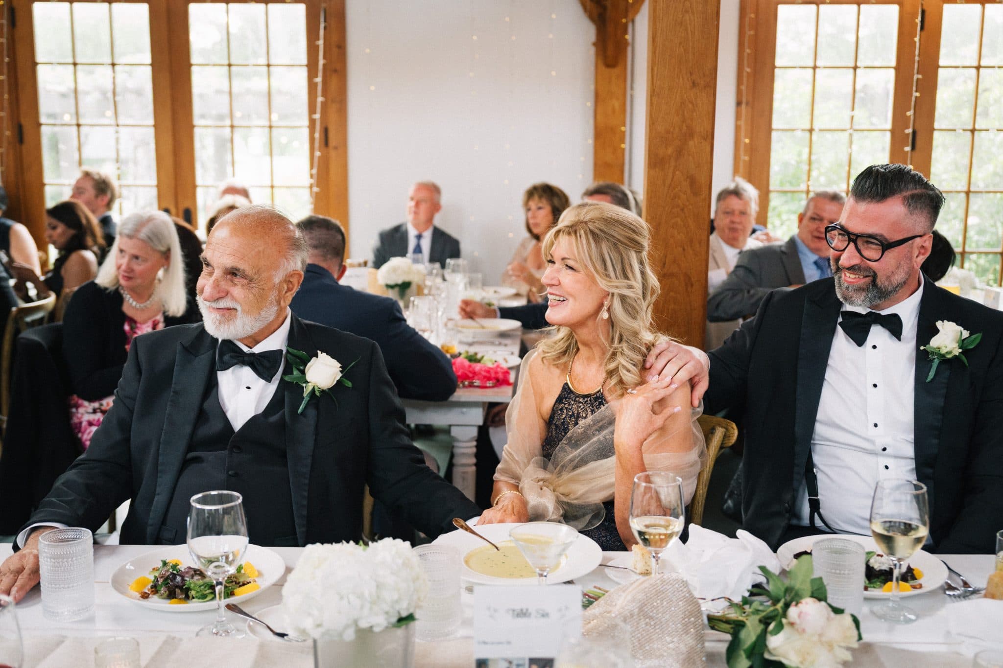 At a wedding reception, three people sit at a table adorned with flowers and food, smiling and enjoying the moment.