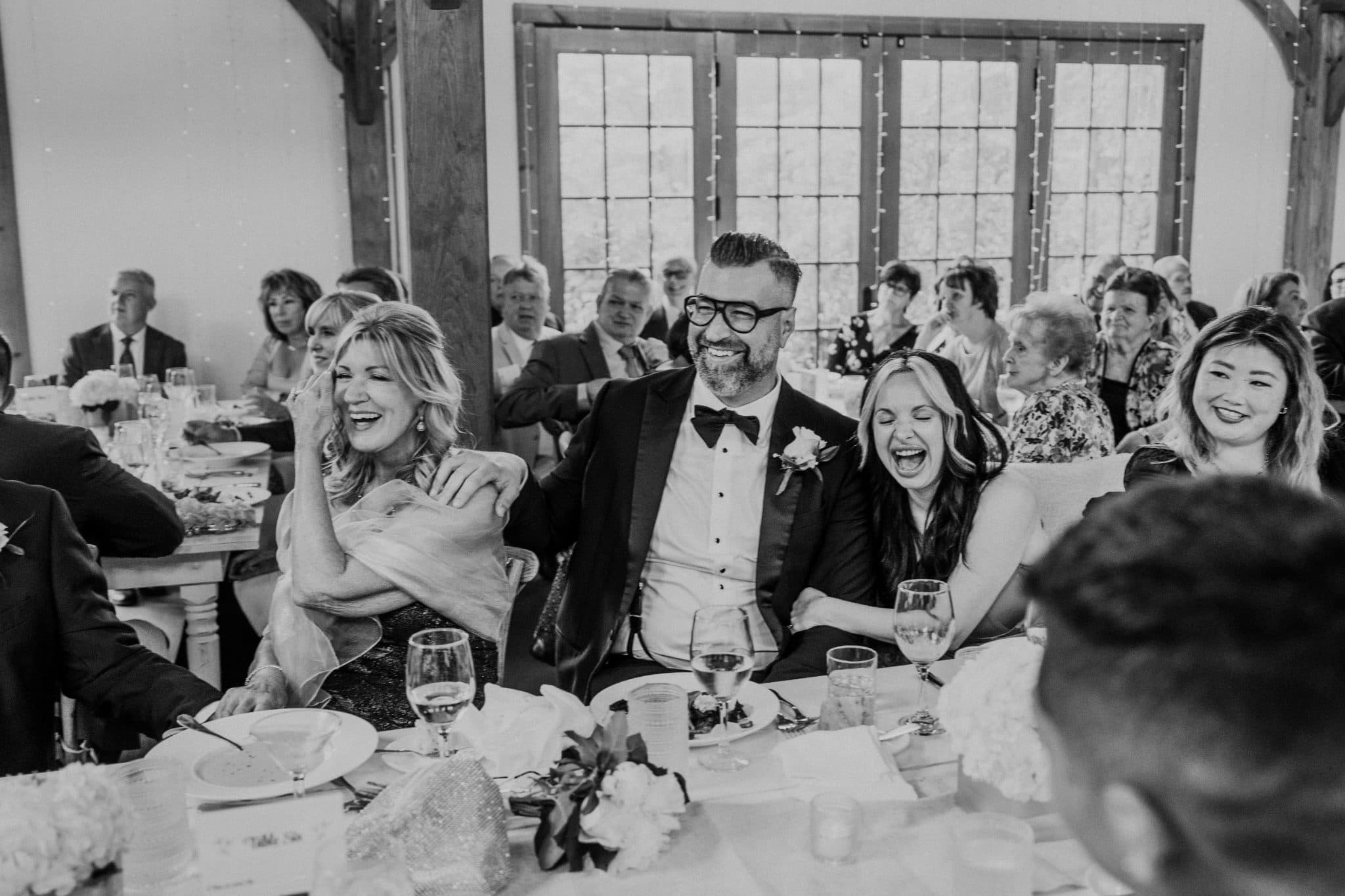 A black-and-white wedding reception scene shows a group of people seated at a table, laughing and enjoying the moment.