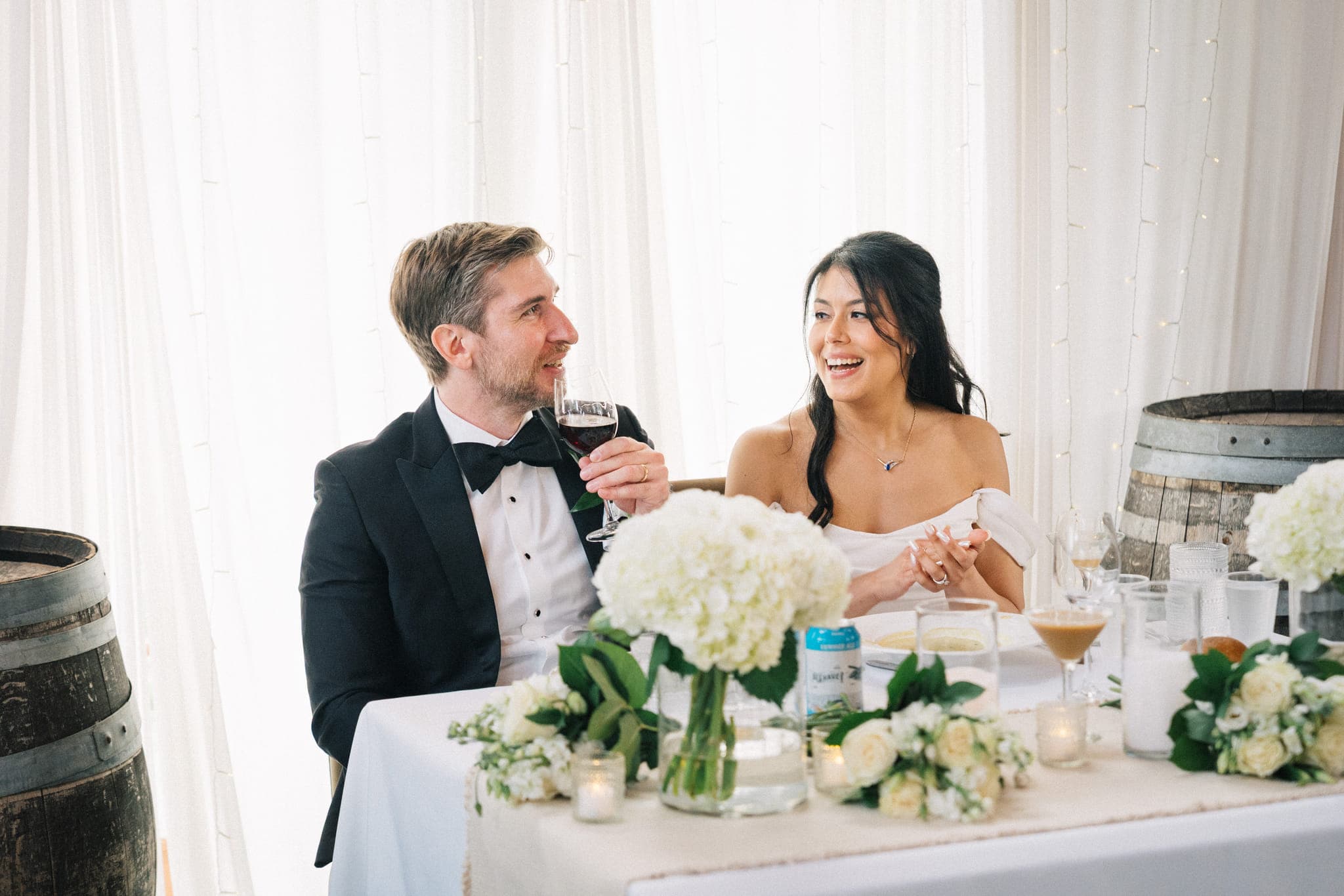 A couple sits at a table adorned with white flowers and wine barrels, sharing a moment of laughter and toasting with wine.