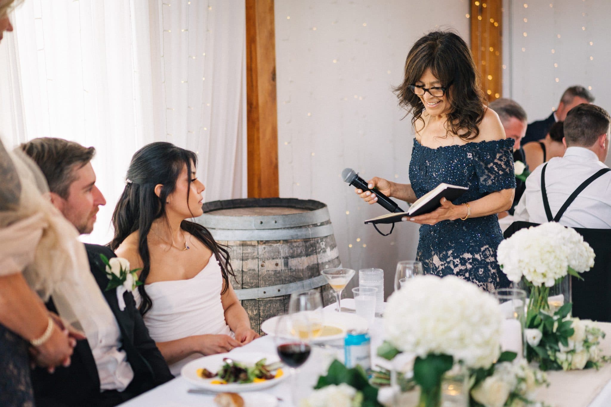 A woman in a blue dress holds a microphone and reads from a book at a wedding reception.