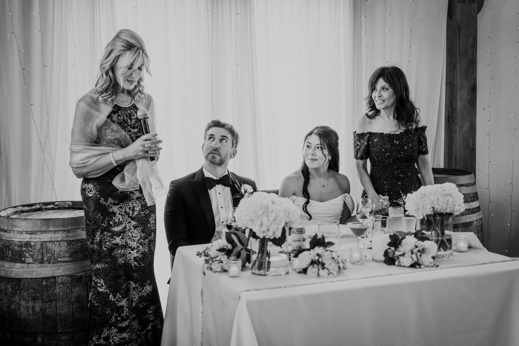 A woman in a patterned dress stands beside a table with a bride and groom, holding a microphone, while another woman in a sequined top looks on.