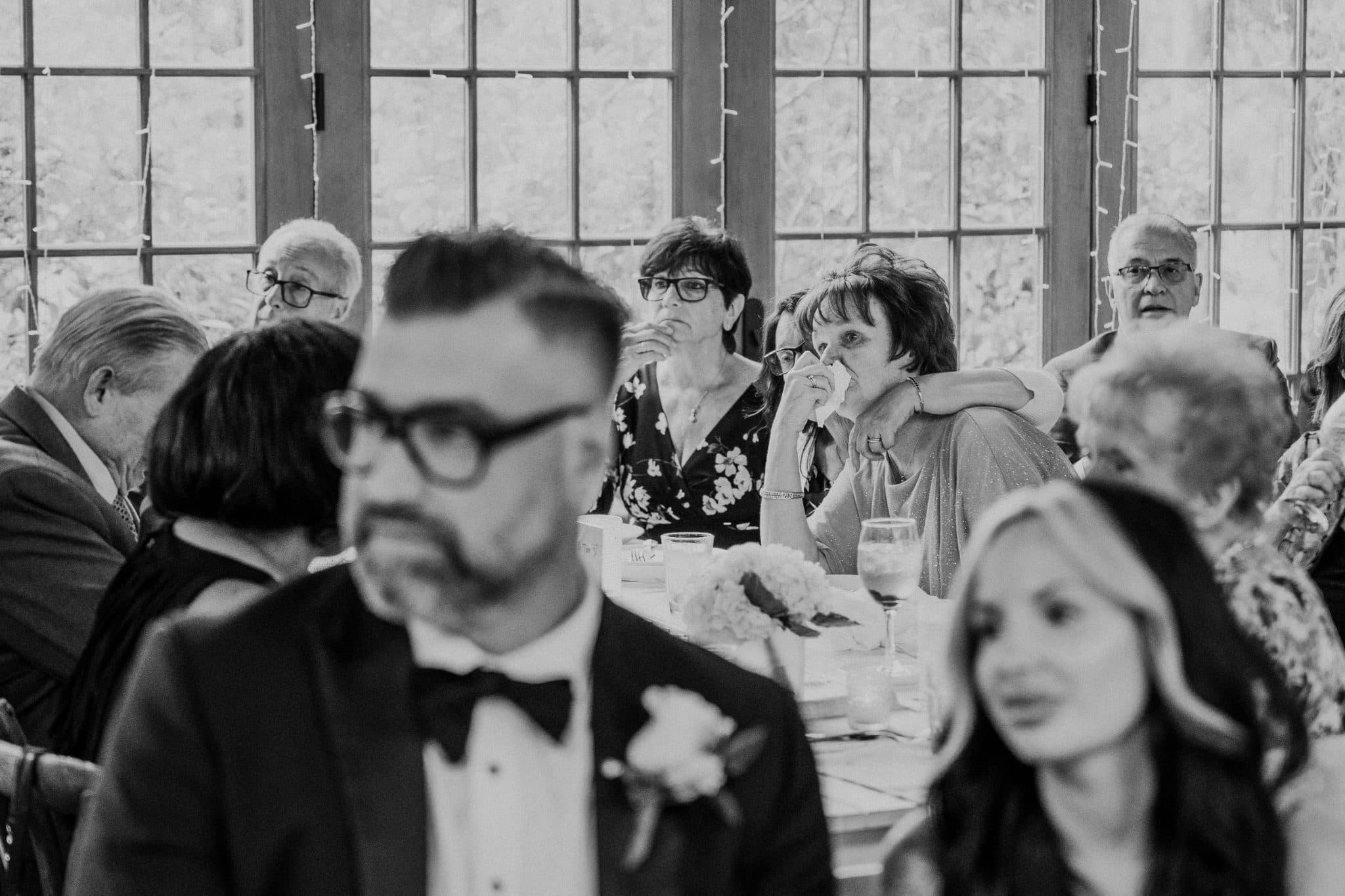 A black-and-white wedding photograph shows a group of people seated at a table, with a man in a tuxedo in the foreground and others in formal attire in the background.