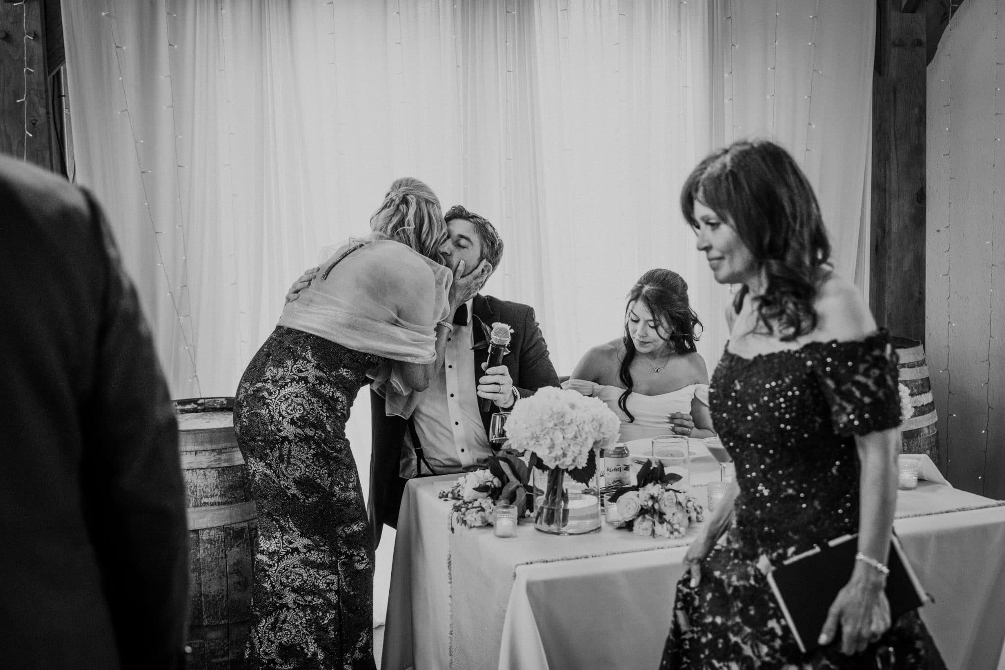 A woman leans over to kiss a man seated at a table with a bride and another woman nearby.