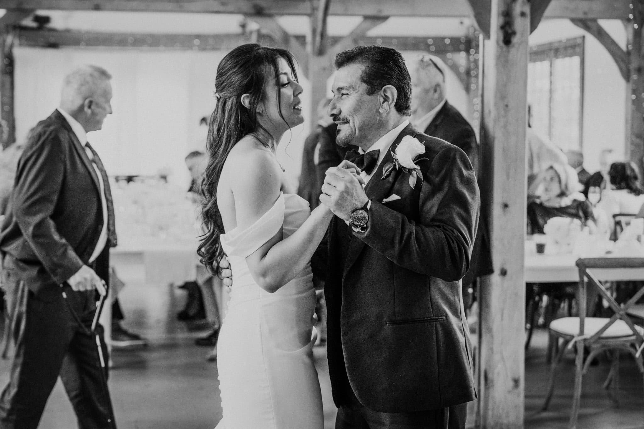 A bride in a white dress dances with a man in a suit adorned with a boutonniere in a barn setting.