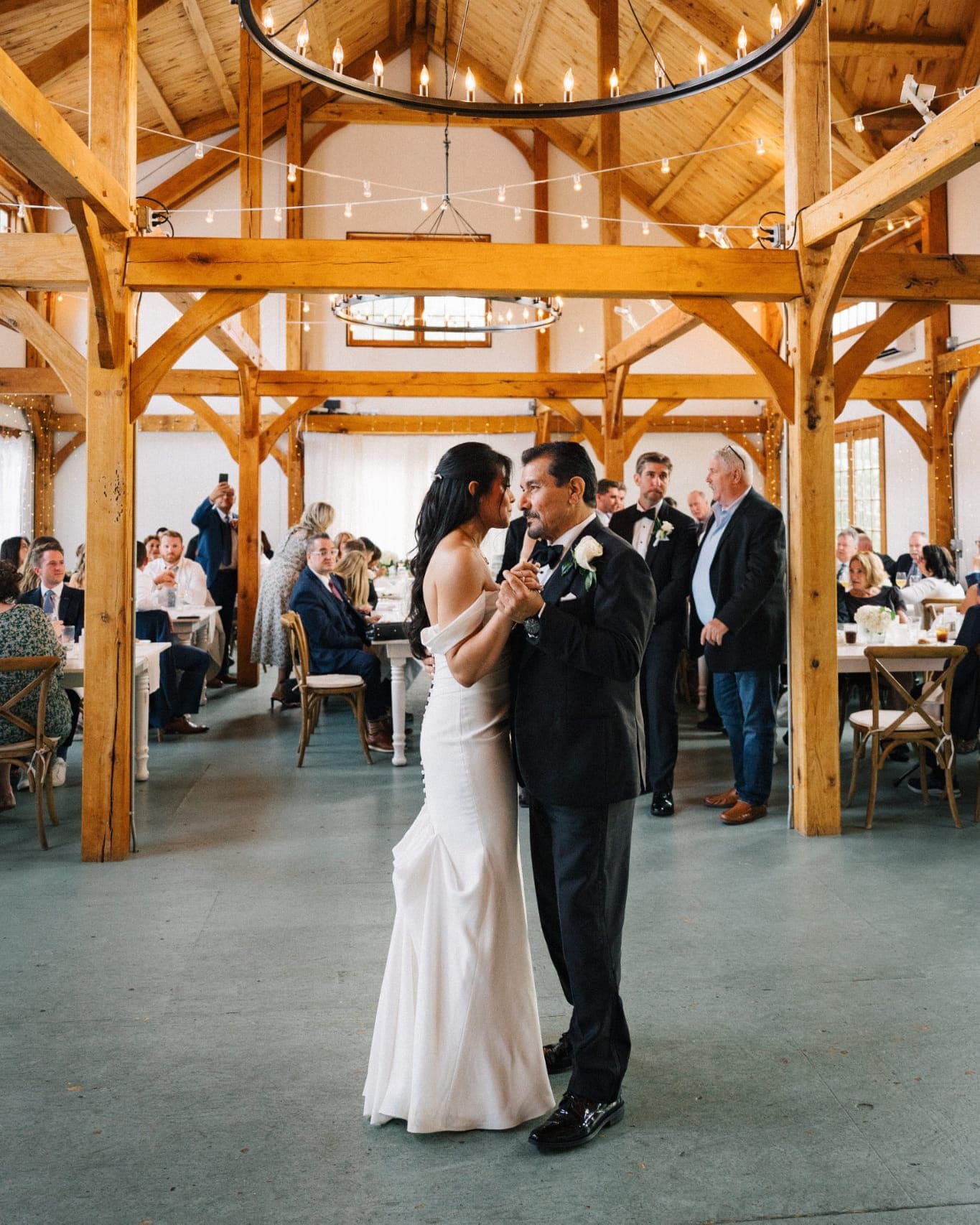 A bride and groom share a dance in a rustic barn with wooden beams and chandeliers.