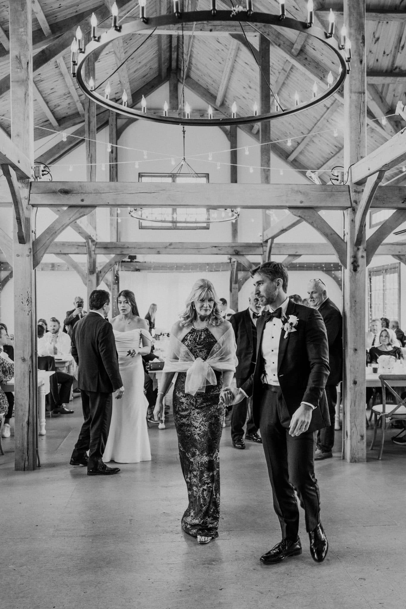 A bride in a white gown walks with her mother in a black and white patterned dress, surrounded by guests in a rustic wooden hall.