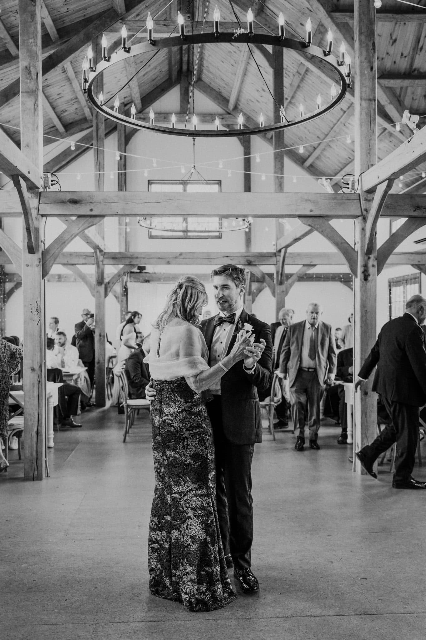 A couple dances under a chandelier in a rustic wooden hall with guests seated at tables in the background.