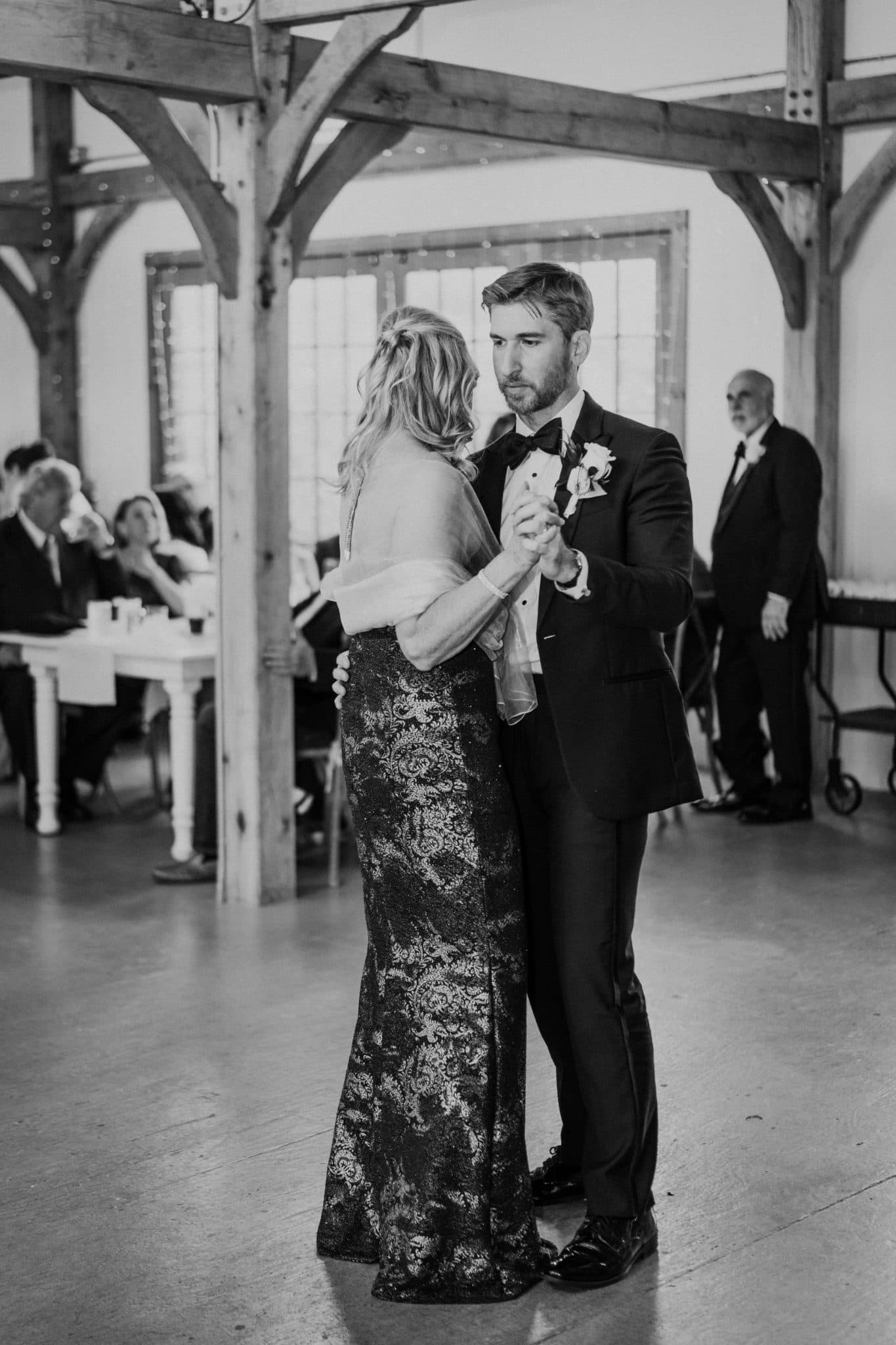 A man and woman in formal attire dance closely in a rustic, wood-beamed room with other guests seated at tables in the background.