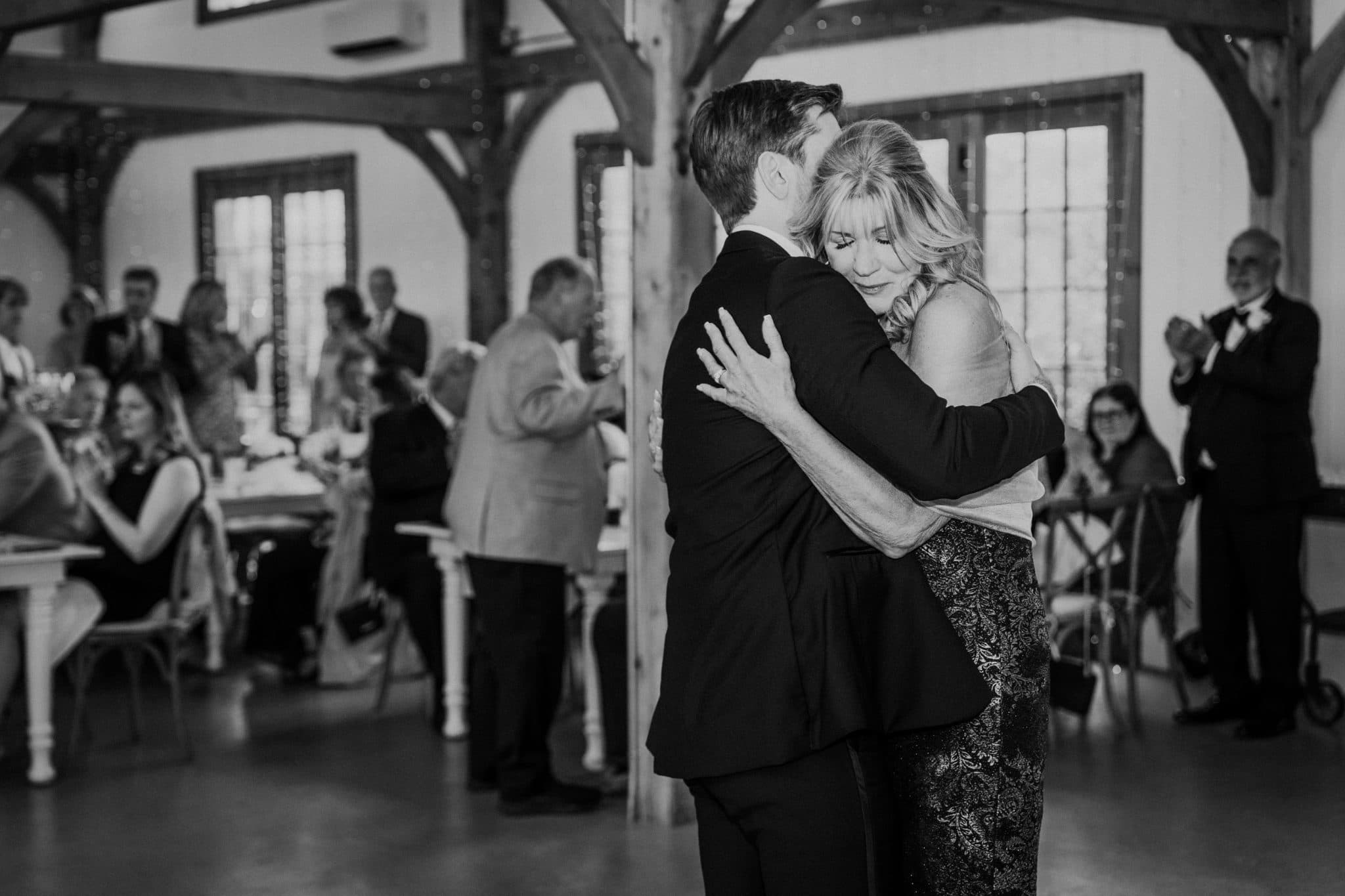 A couple embraces warmly in a rustic indoor setting, surrounded by seated guests.