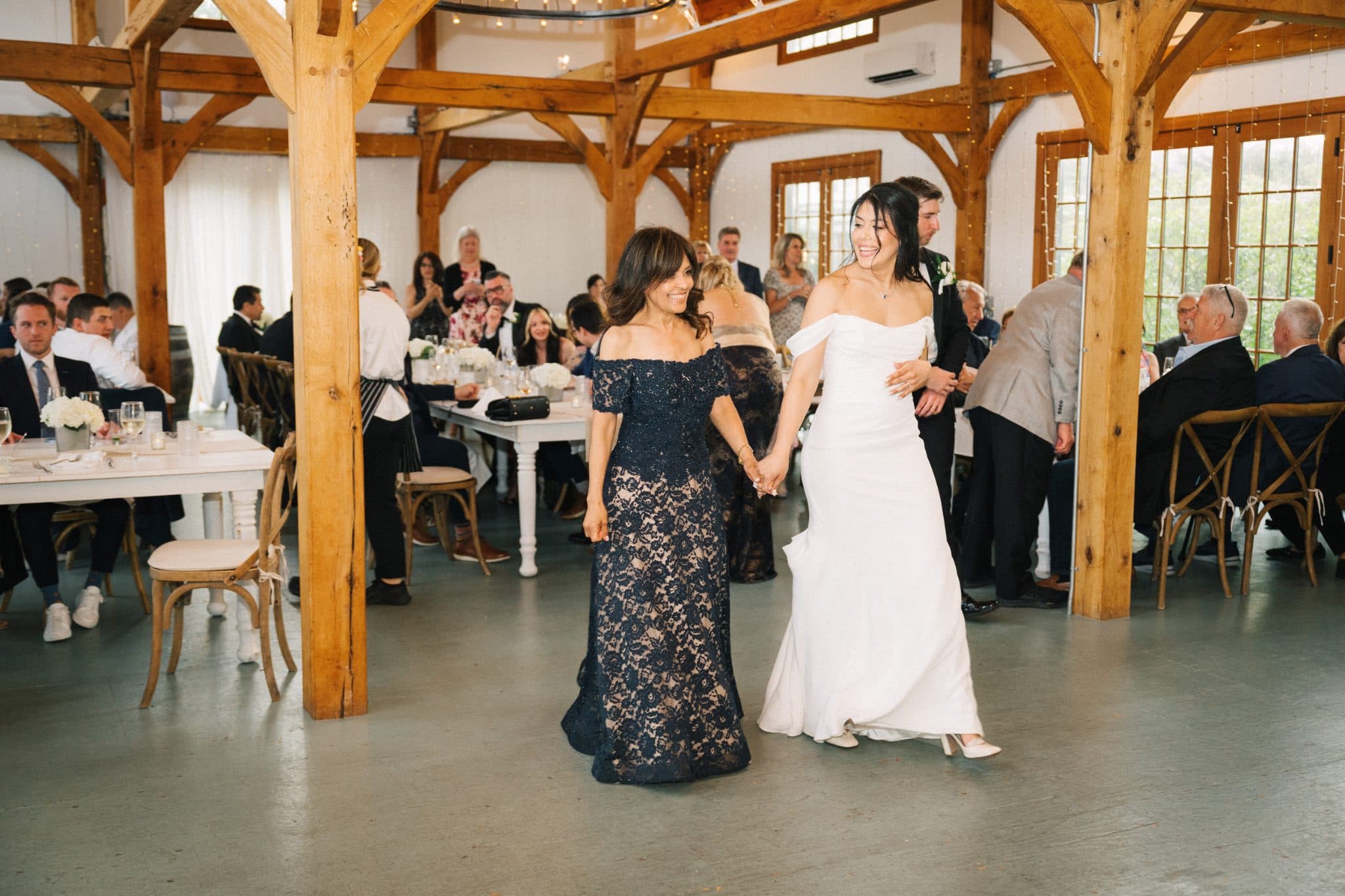 A bride in a white dress dances with a woman in a dark blue lace dress in a wooden barn with guests seated at tables.