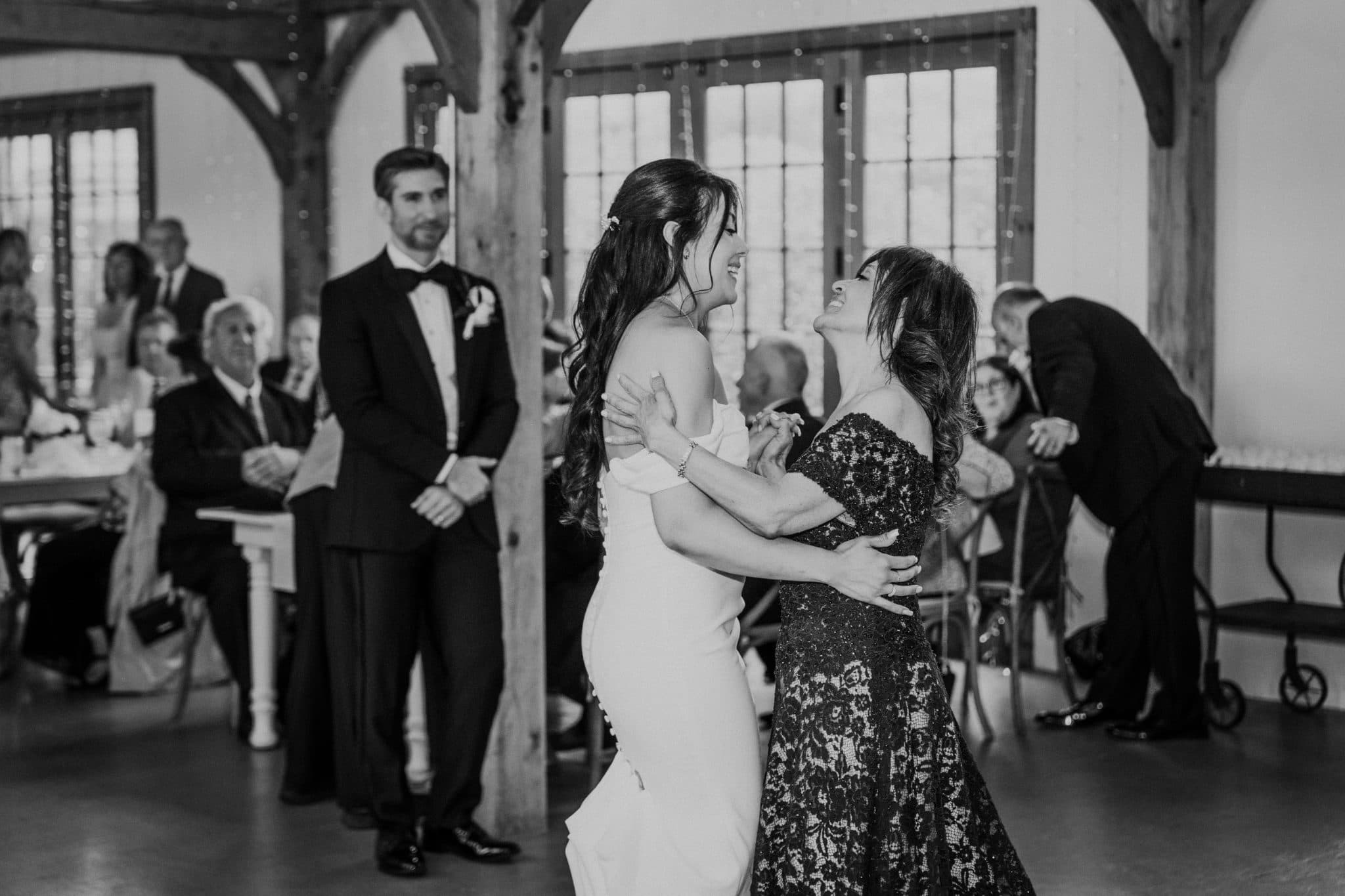 A bride in a white dress embraces another woman in a black lace dress in a room with wooden beams and seated guests.