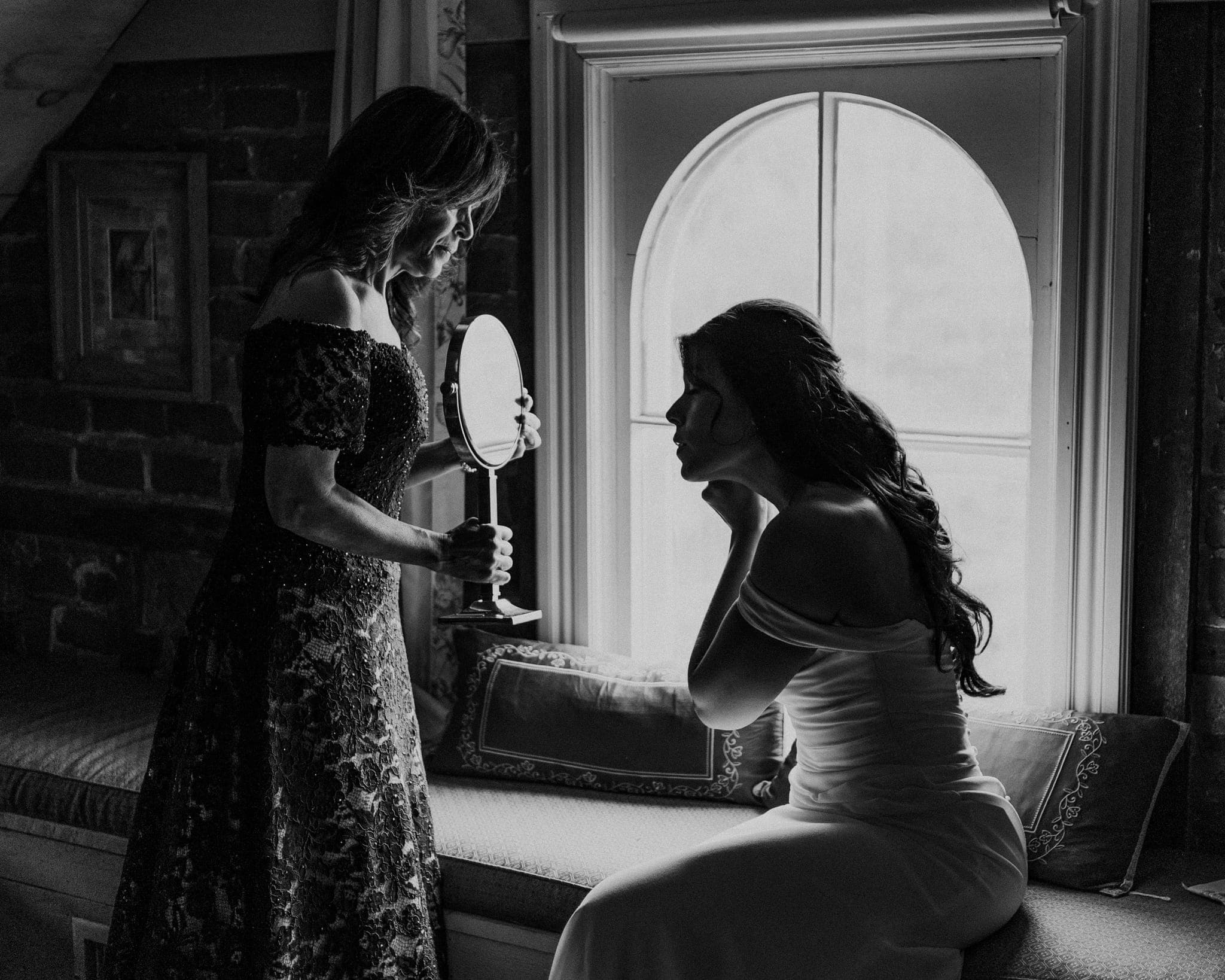 A woman in a lace dress holds a mirror while another woman in a white dress sits on a cushioned bench, looking at herself in the mirror.