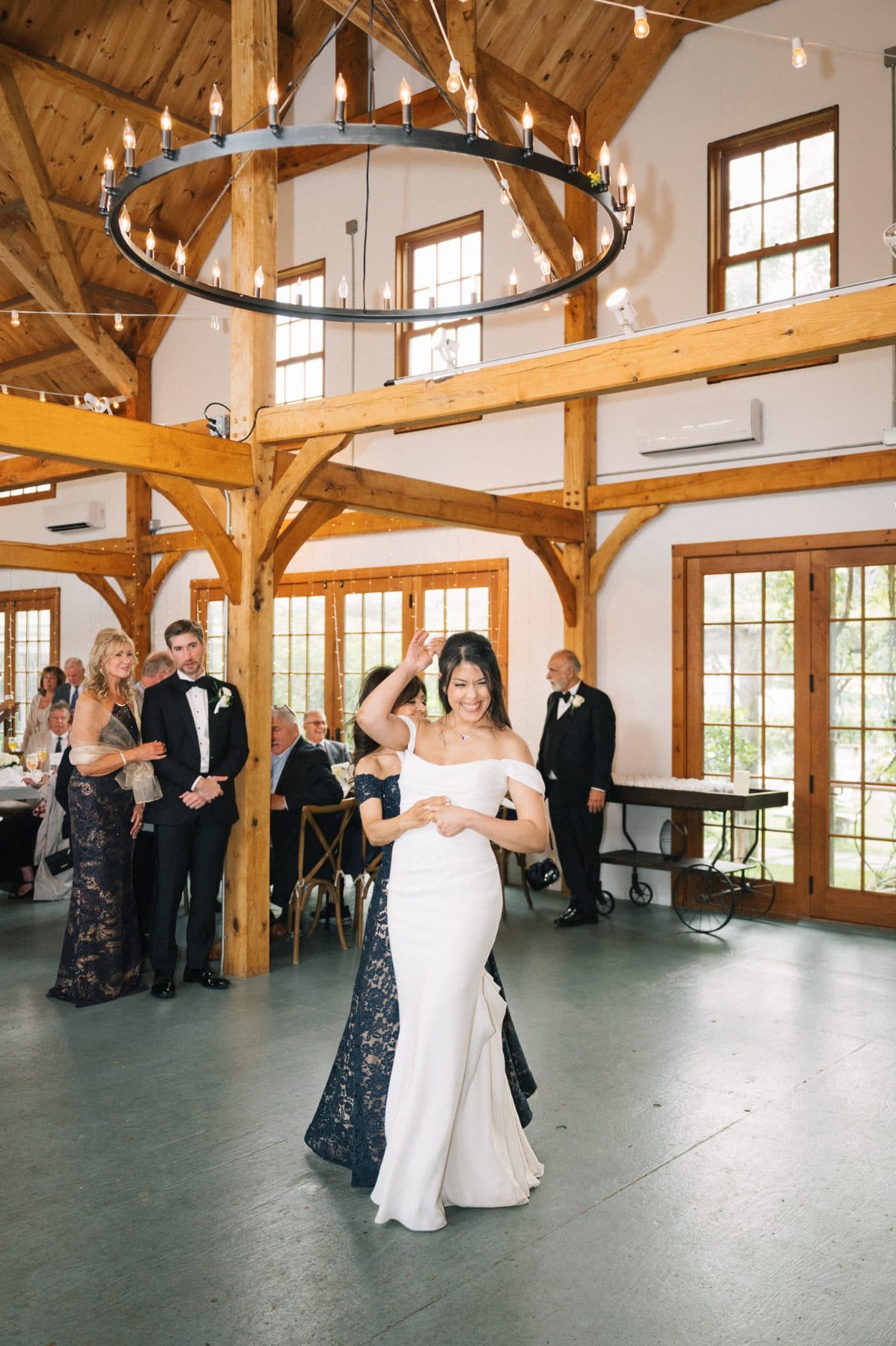 A bride in a white gown dances joyfully in a rustic wooden hall with guests watching.