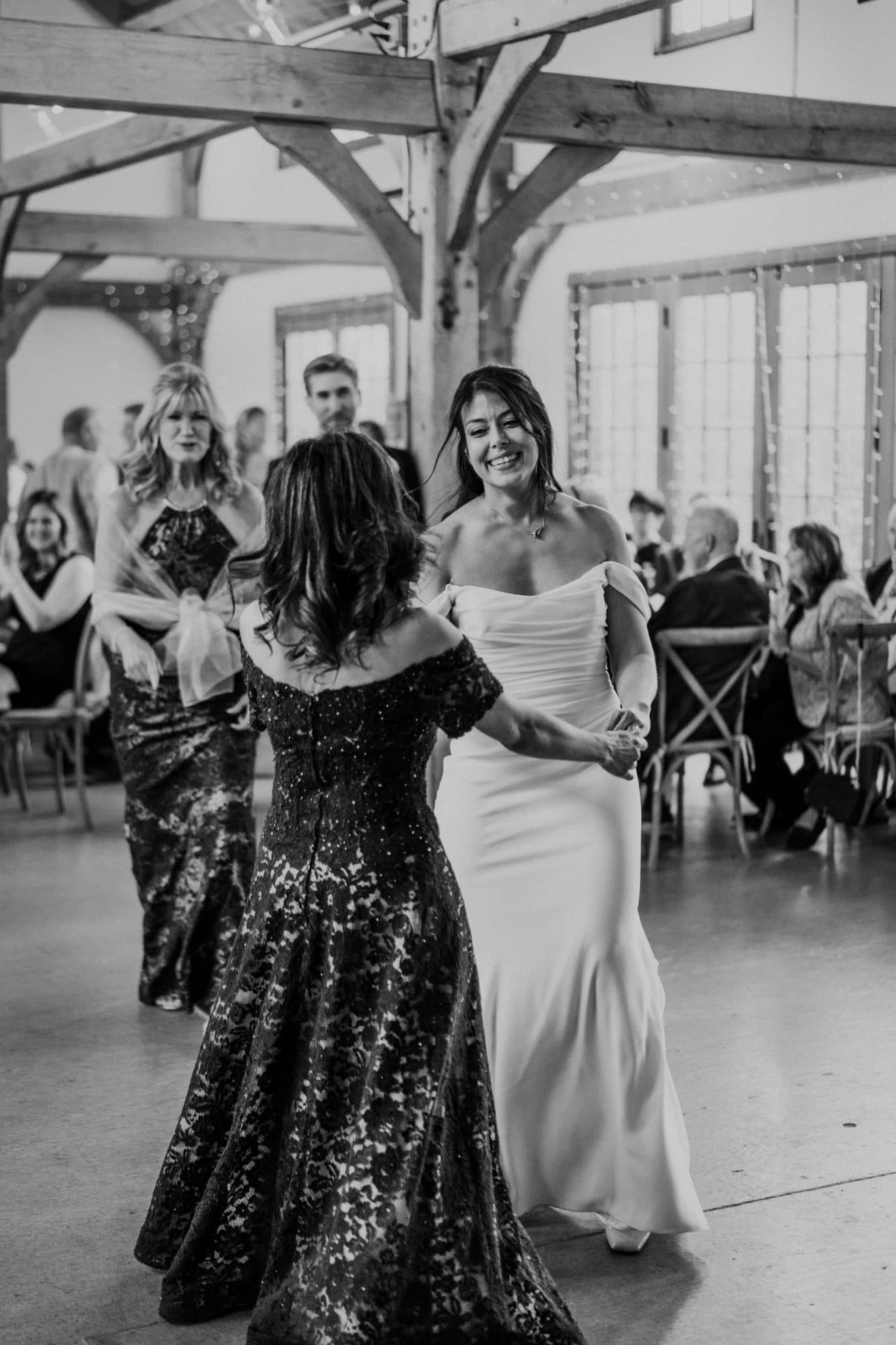 A bride in a white gown dances with a woman in a dark, patterned dress in a barn-style venue.