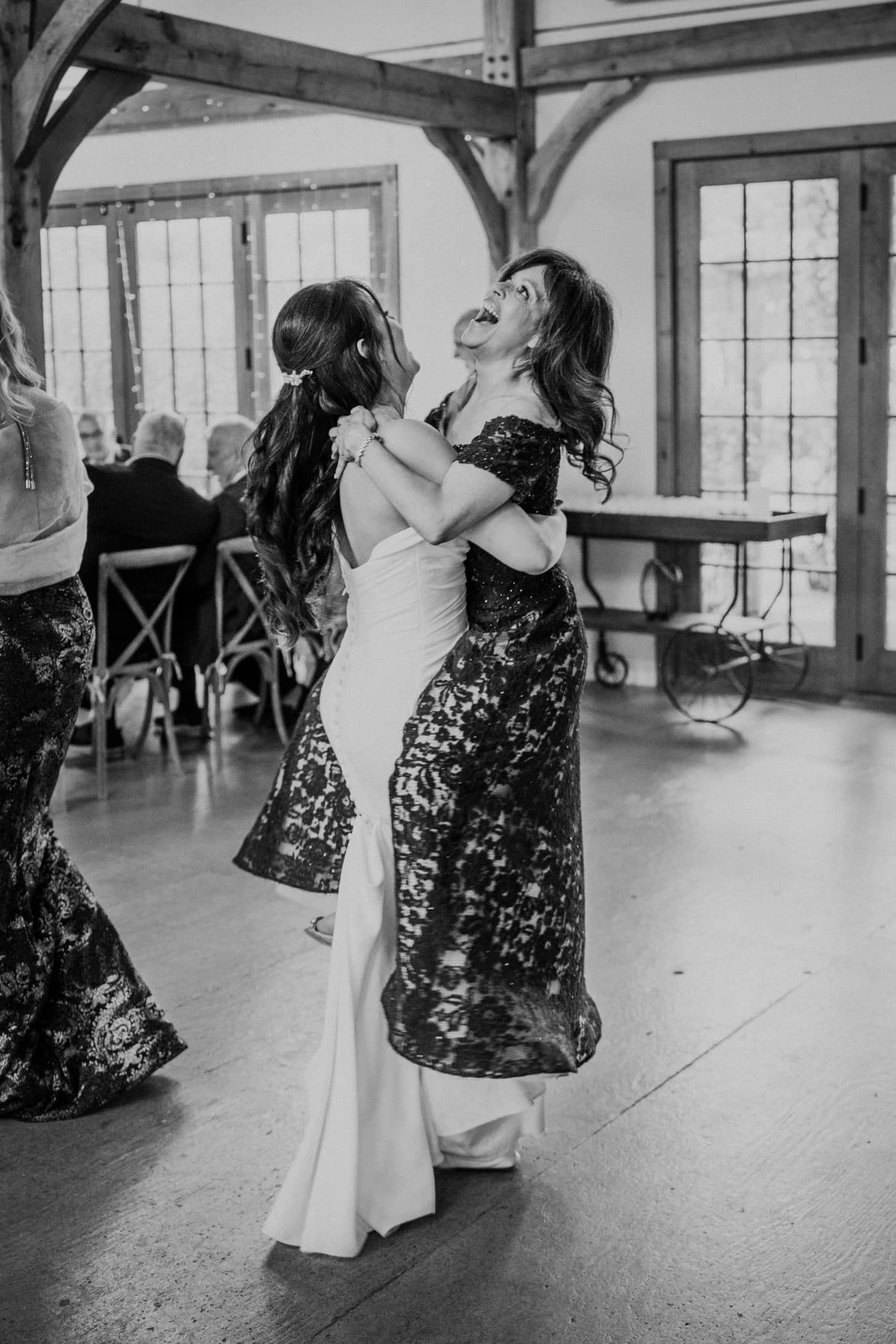A bride in a white dress dances with a guest in a black floral dress in a barn-style venue.