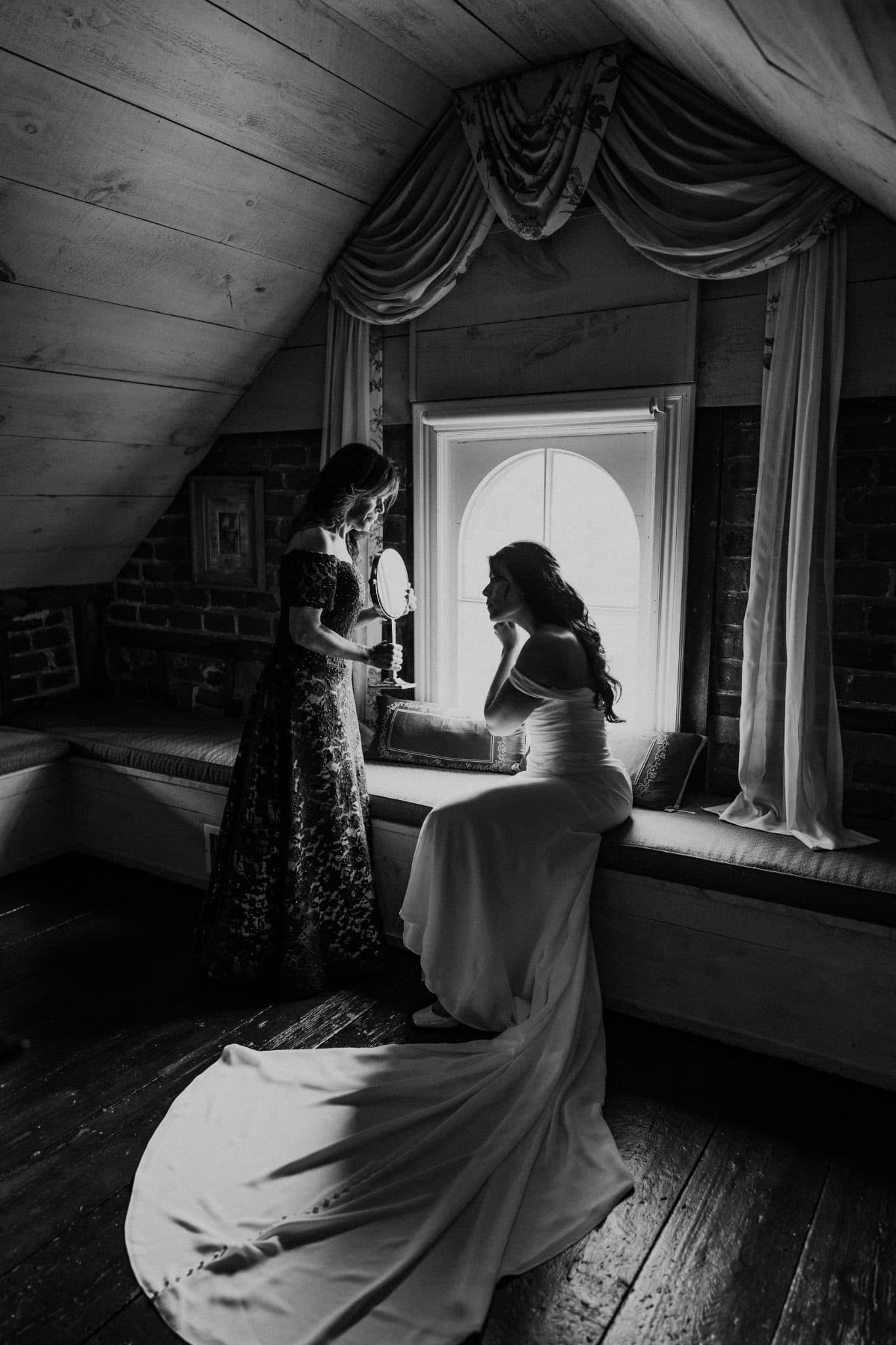 A bride in a flowing dress sits by a window, holding a mirror as another woman stands beside her.
