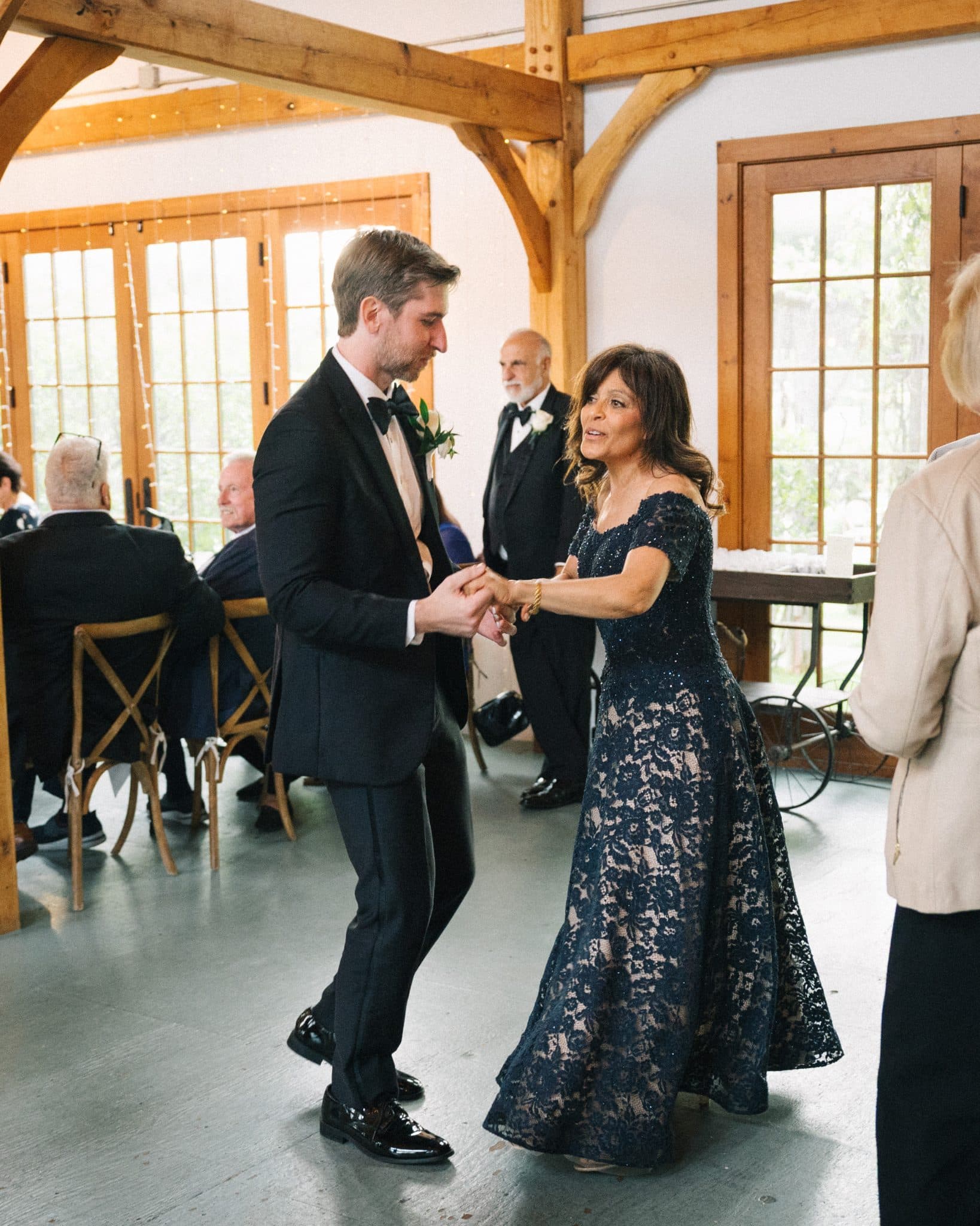 A man in a tuxedo dances with a woman in a lace dress in a room with wooden beams and large windows.