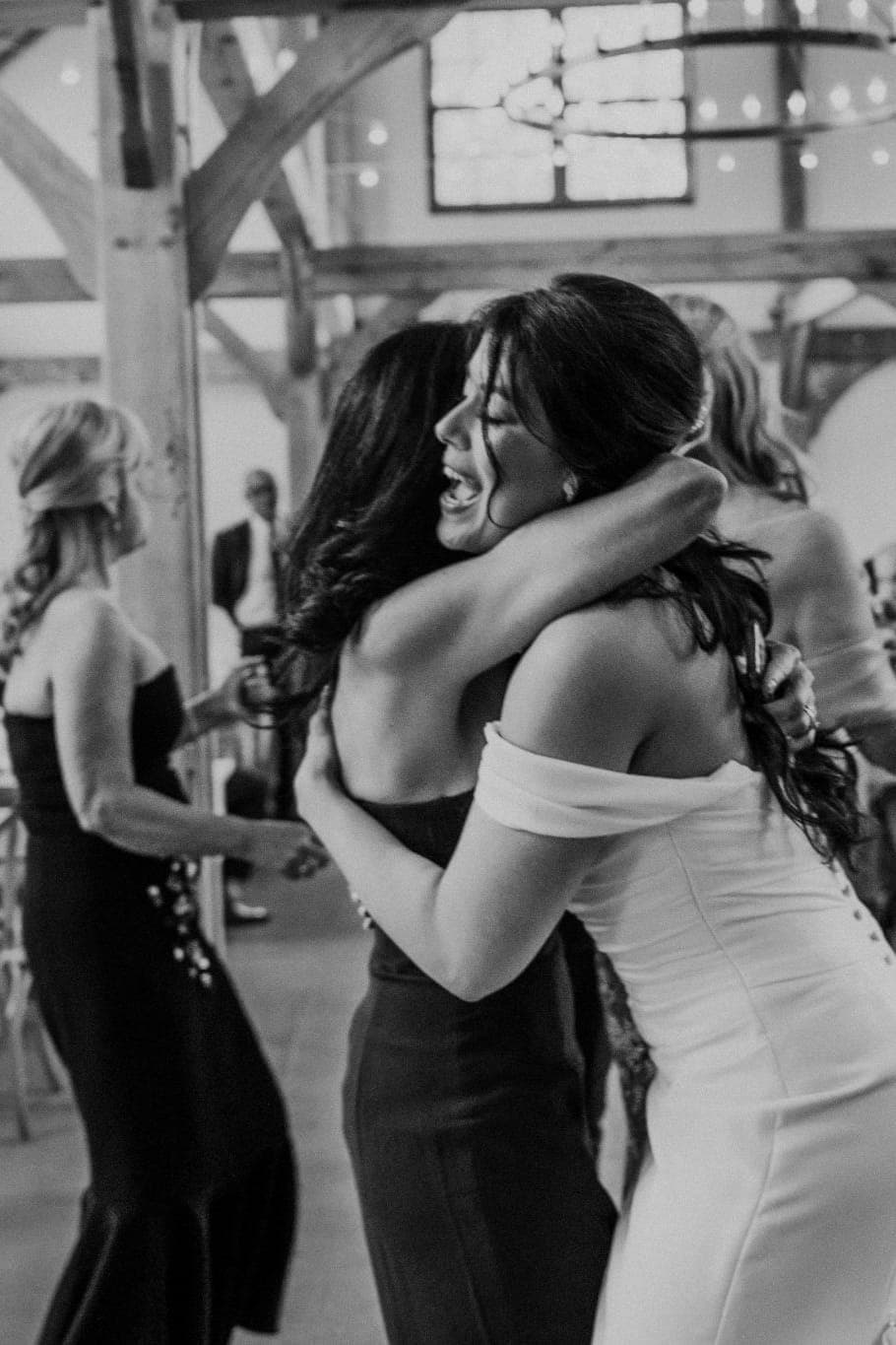 A bride in a white dress embraces another woman in a dark dress in a barn-style wedding reception.