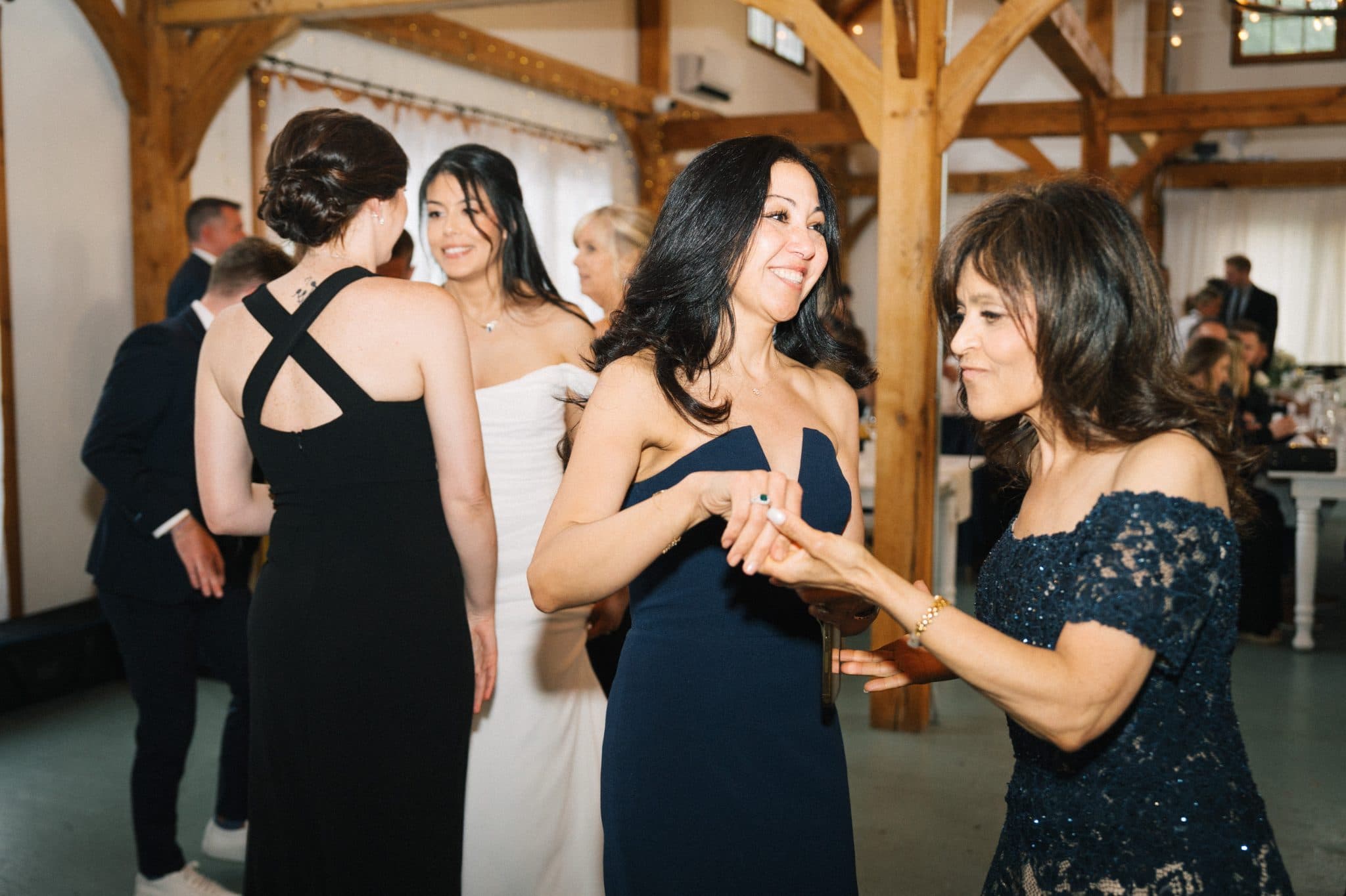 A woman in a dark blue dress dances with another woman in a lace dress at a wedding reception.