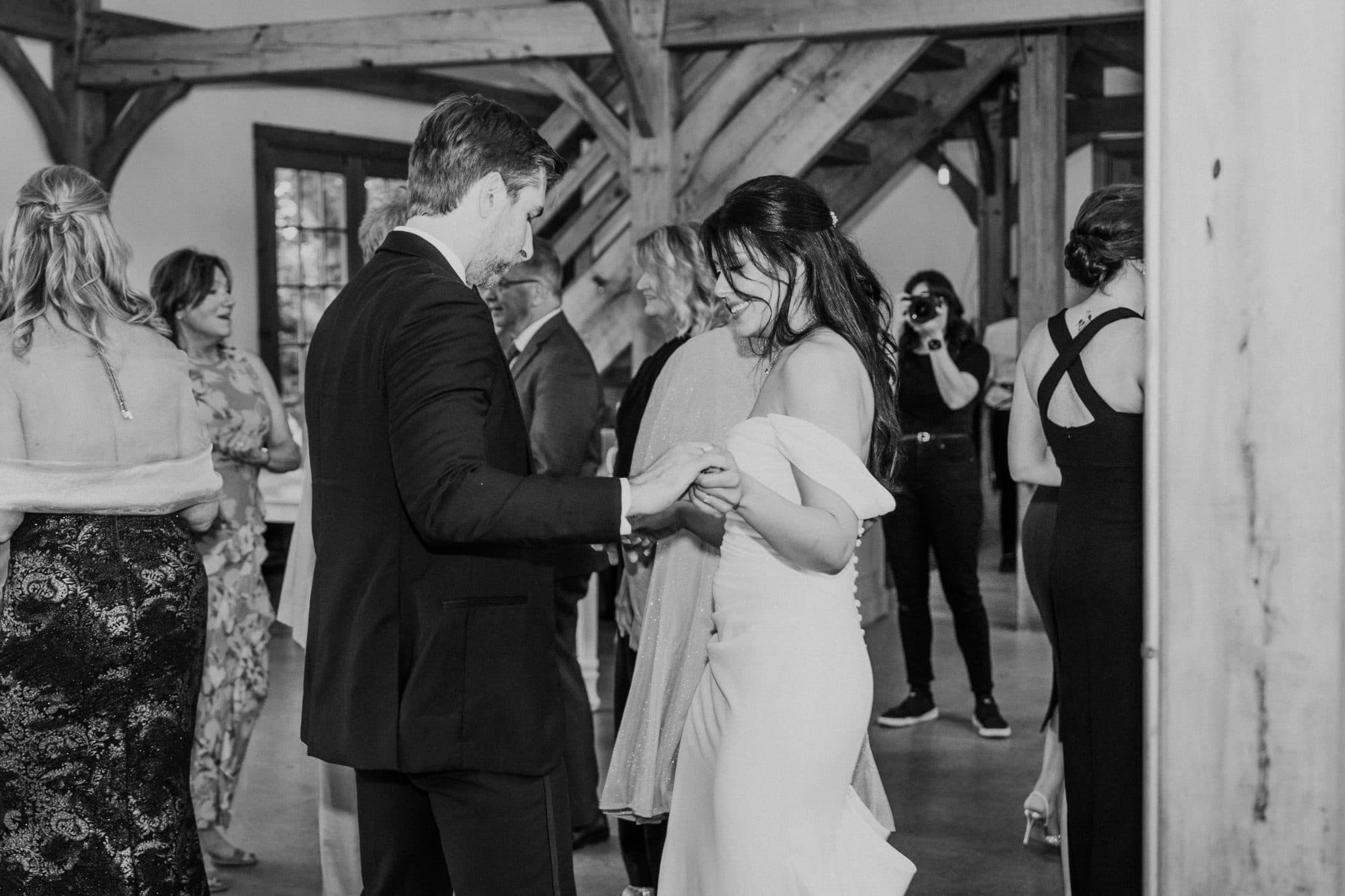 A couple dances closely in a rustic barn setting, surrounded by guests and a photographer.