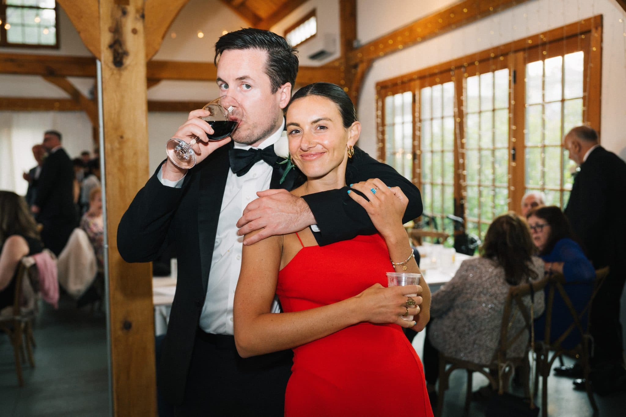 A man in a tuxedo and a woman in a red dress embrace, with the man drinking from a glass.
