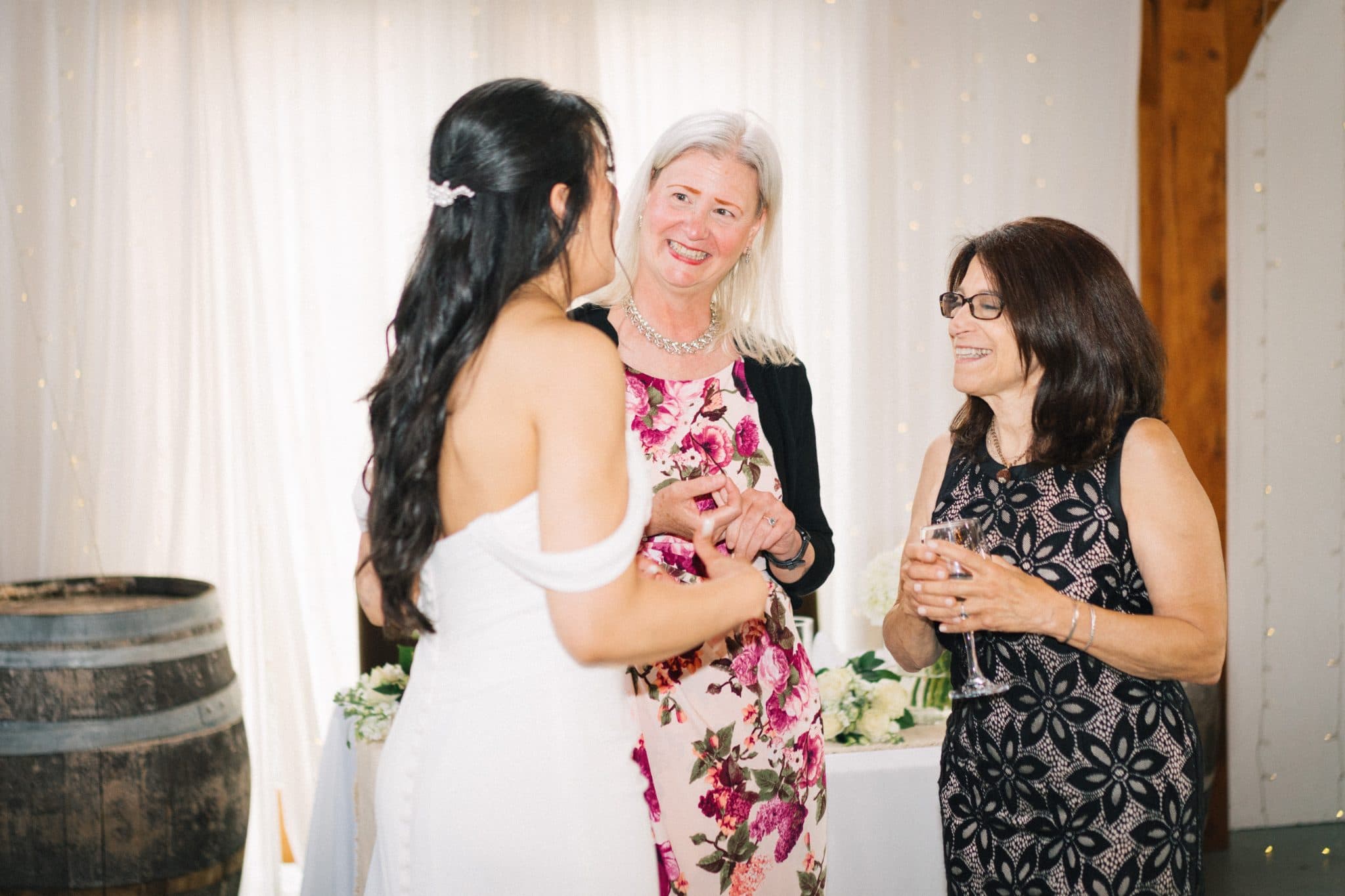 A bride in a white dress interacts with two women in floral dresses, one holding a glass, against a backdrop of white curtains and a wooden barrel.