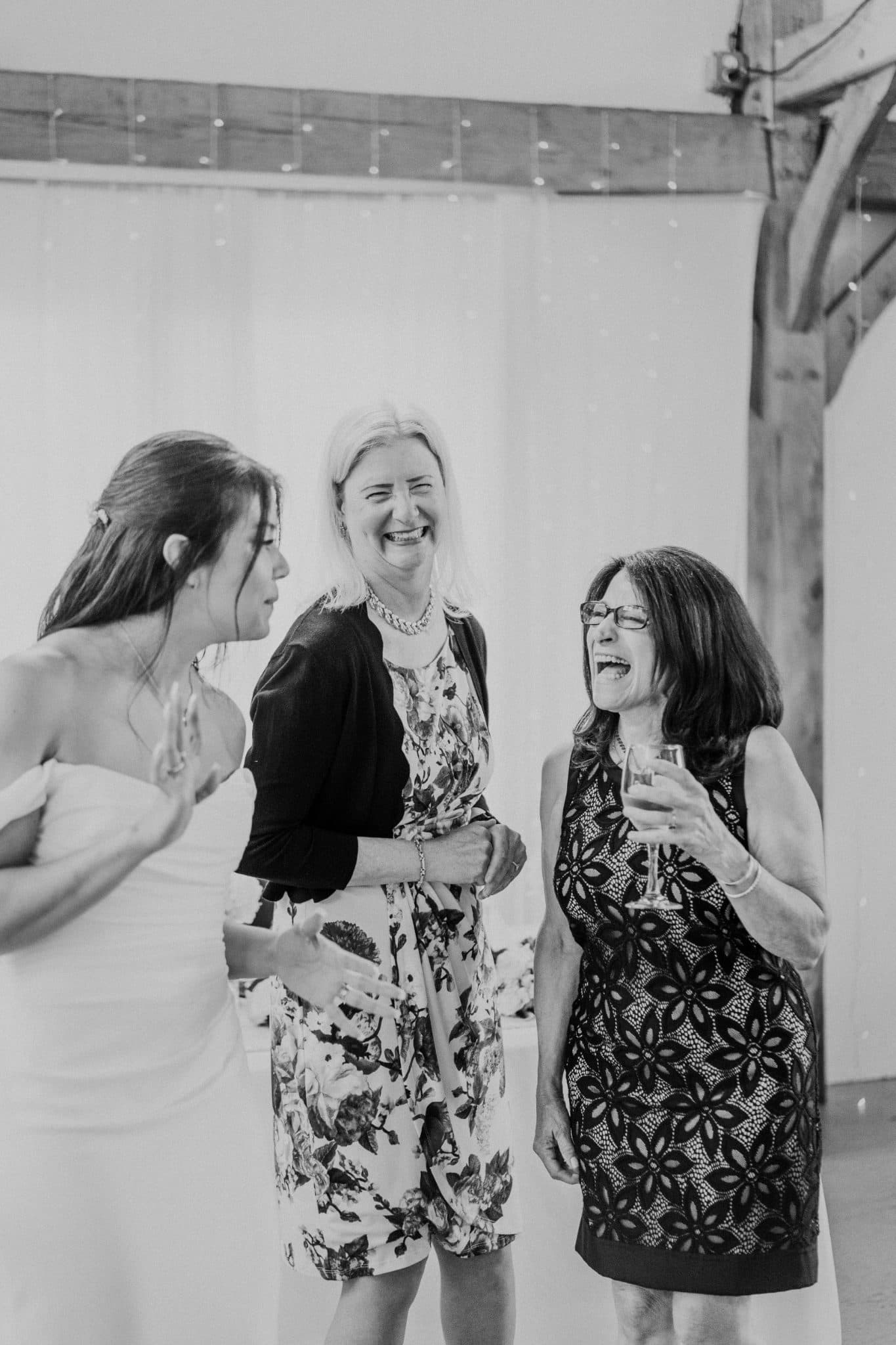 Three women in dresses are laughing and holding drinks in a room with a white wall and wooden beams.