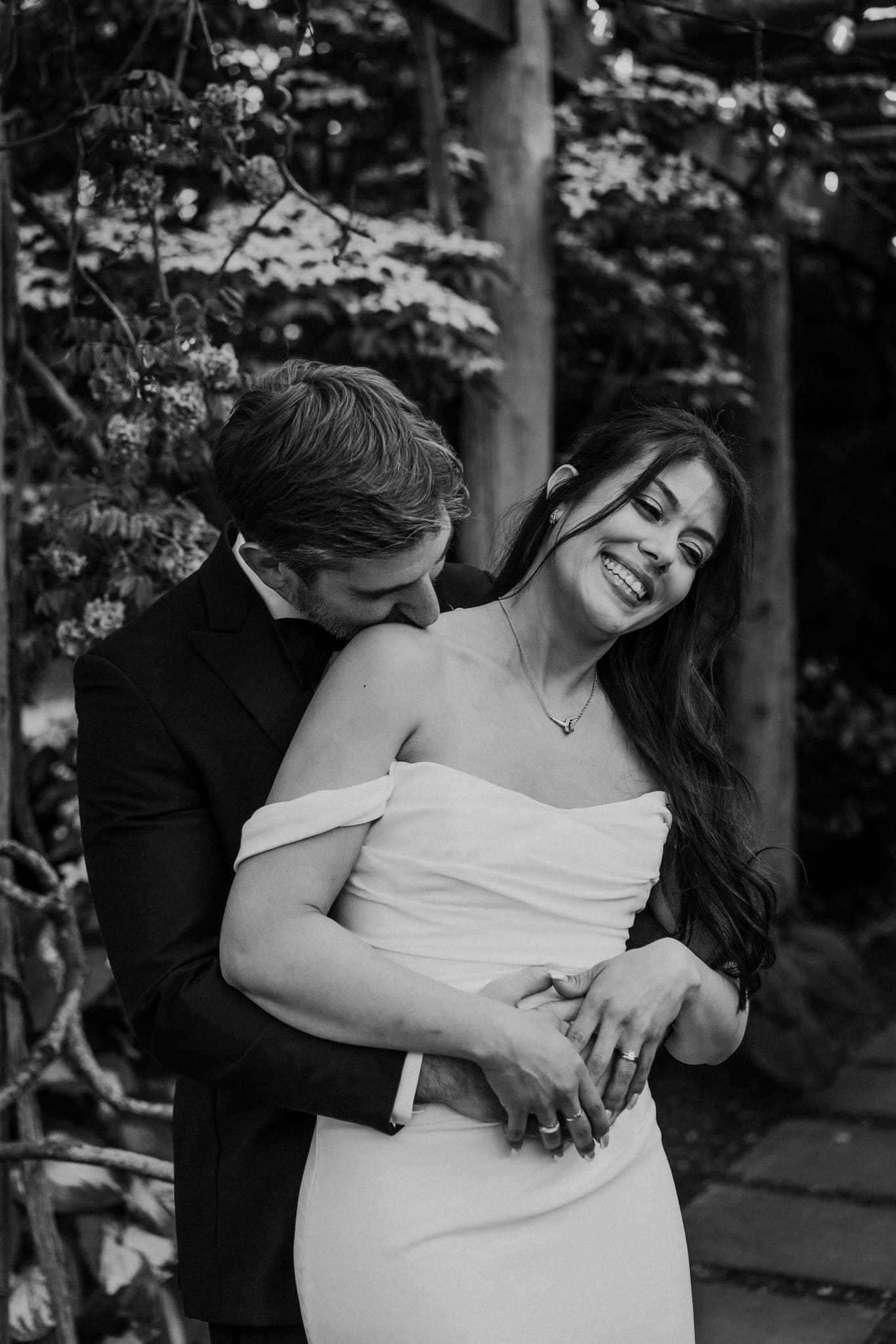 A couple embraces in a garden, the woman in a strapless dress and the man in a suit.