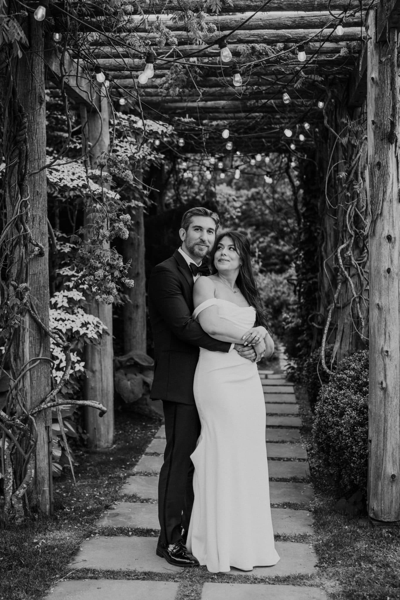 A bride and groom stand embraced under a wooden pergola adorned with string lights.